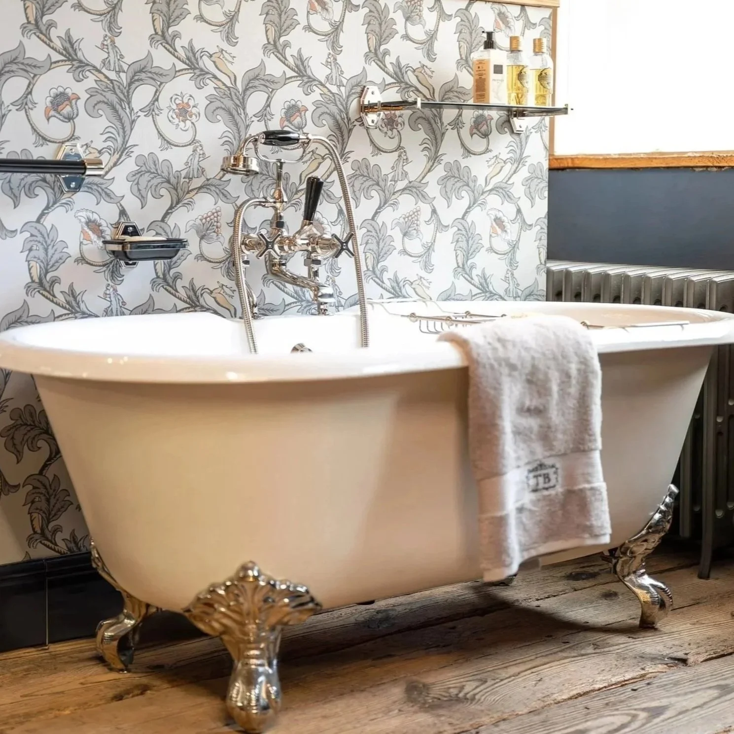 Eine weiße Badewanne im Vintage-Stil mit silbernen, verschnörkelten Beinen, einer Handbrause und einem Wasserhahn. Es hängen Handtücher an der Seite, und in der Nähe gibt es Regale mit Flaschen. Die Wand ist mit Blumendruckem tapeziert, und der Boden