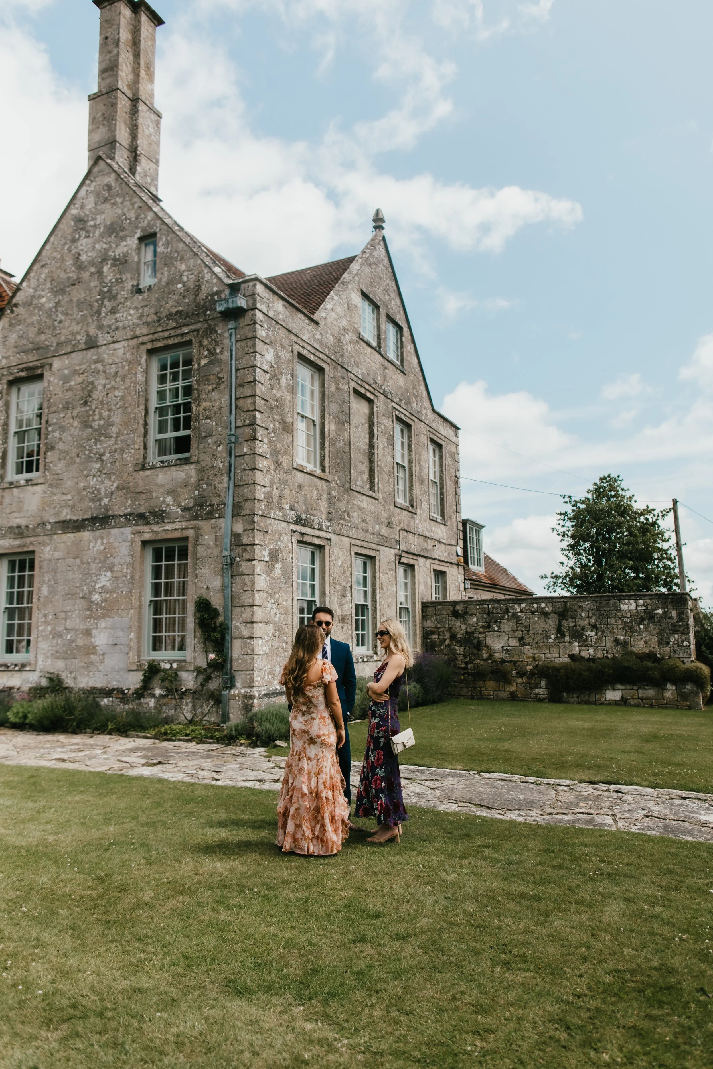 Three people, two women and a man, standing on the lawn in front of Hatch House with large windows and a steep roof, engaged in conversation on a sunny day with scattered clouds.