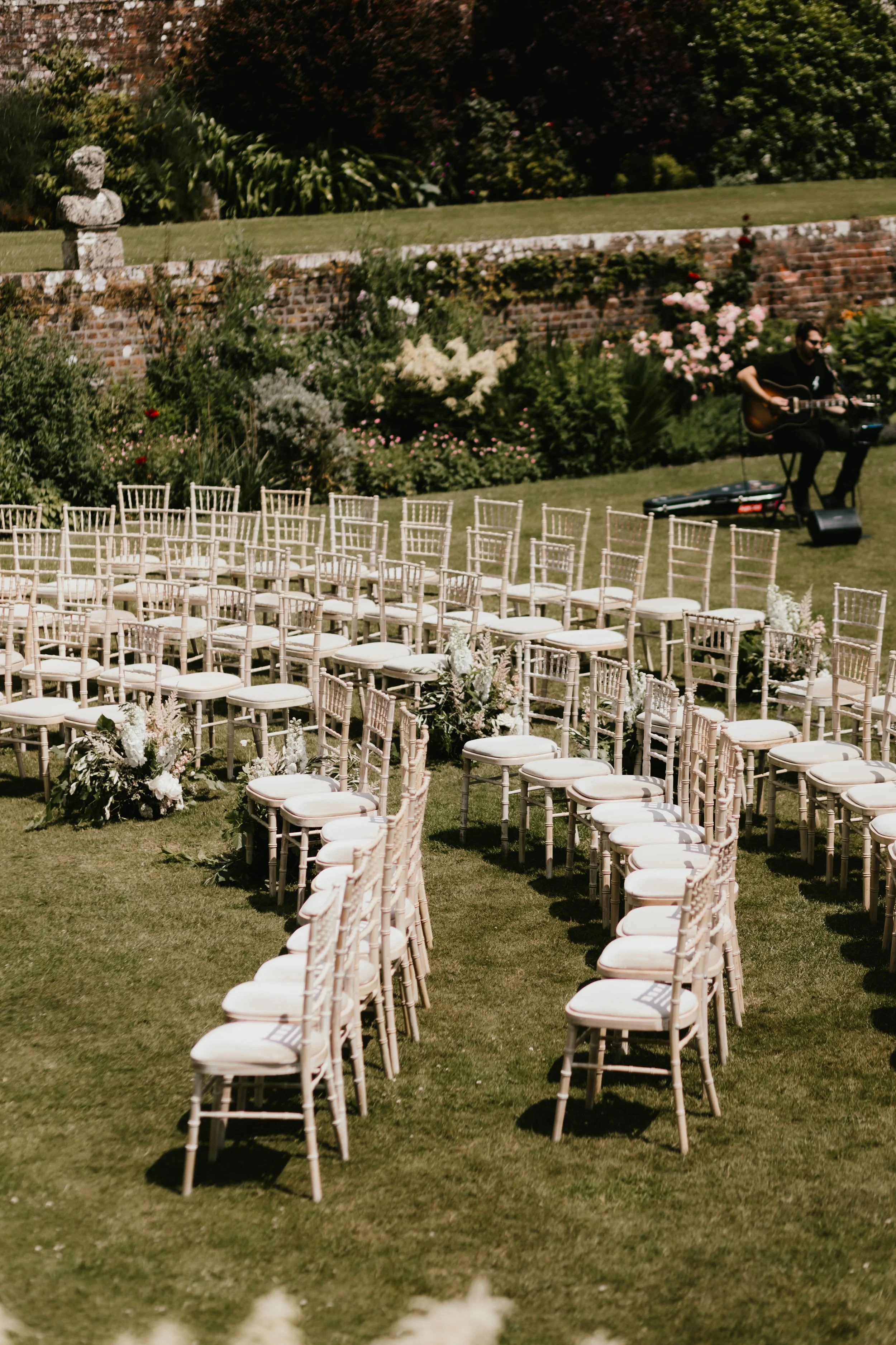 Wedding ceremony setup outdoors with rows of white chairs on green grass, decorated with floral arrangements, at Hatch House. A musician is playing guitar in the background.