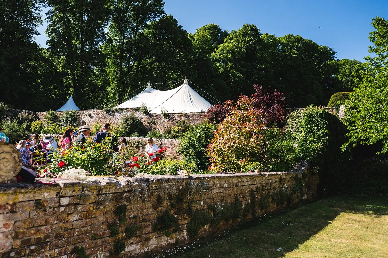 People gathered outdoors in a lush garden with blooming flowers, stone walls, and tents in the background under a clear blue sky.