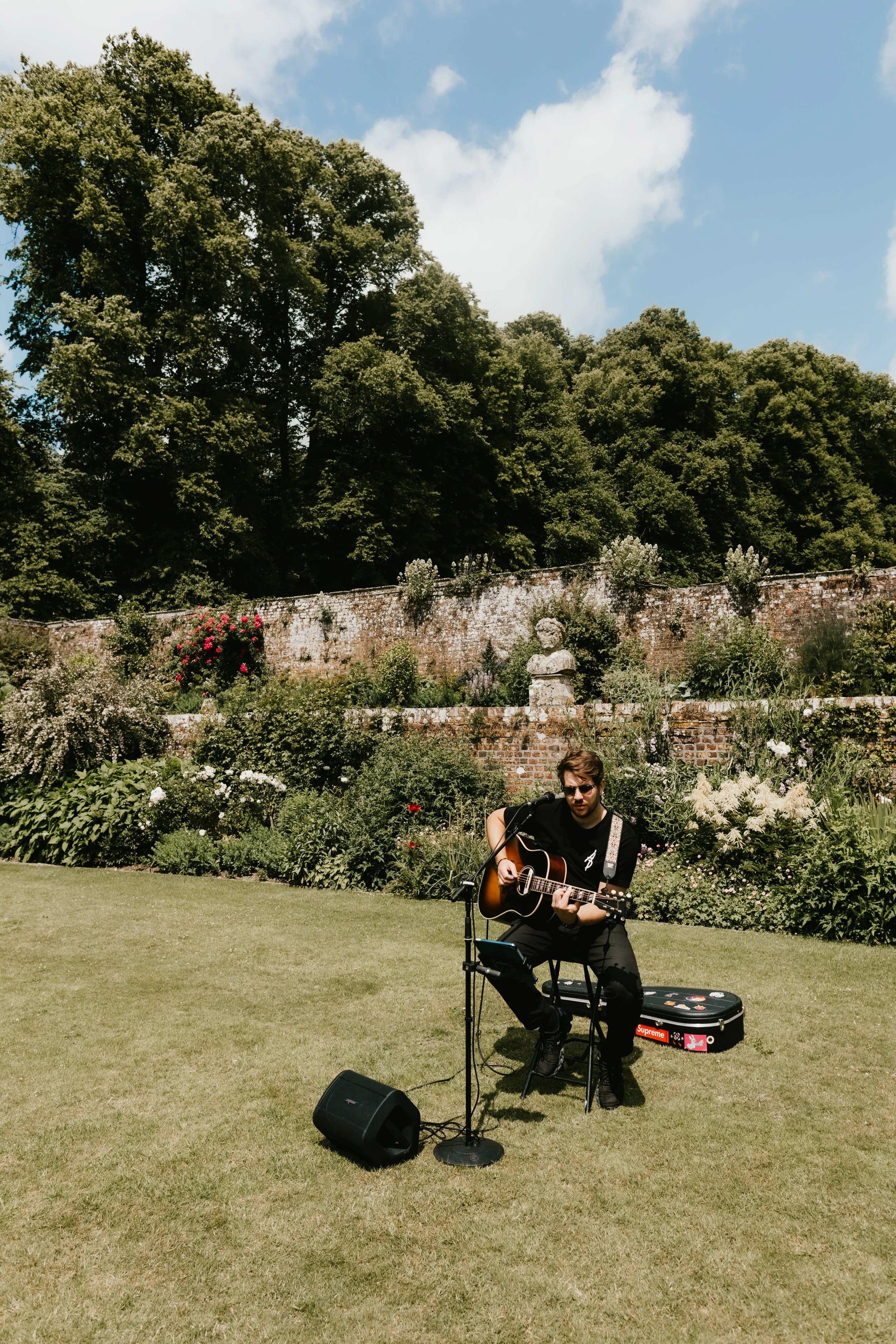 A man with sunglasses playing acoustic guitar outdoors on the lawn at Hatch House, with a garden and brick wall behind him, under a partly cloudy sky.