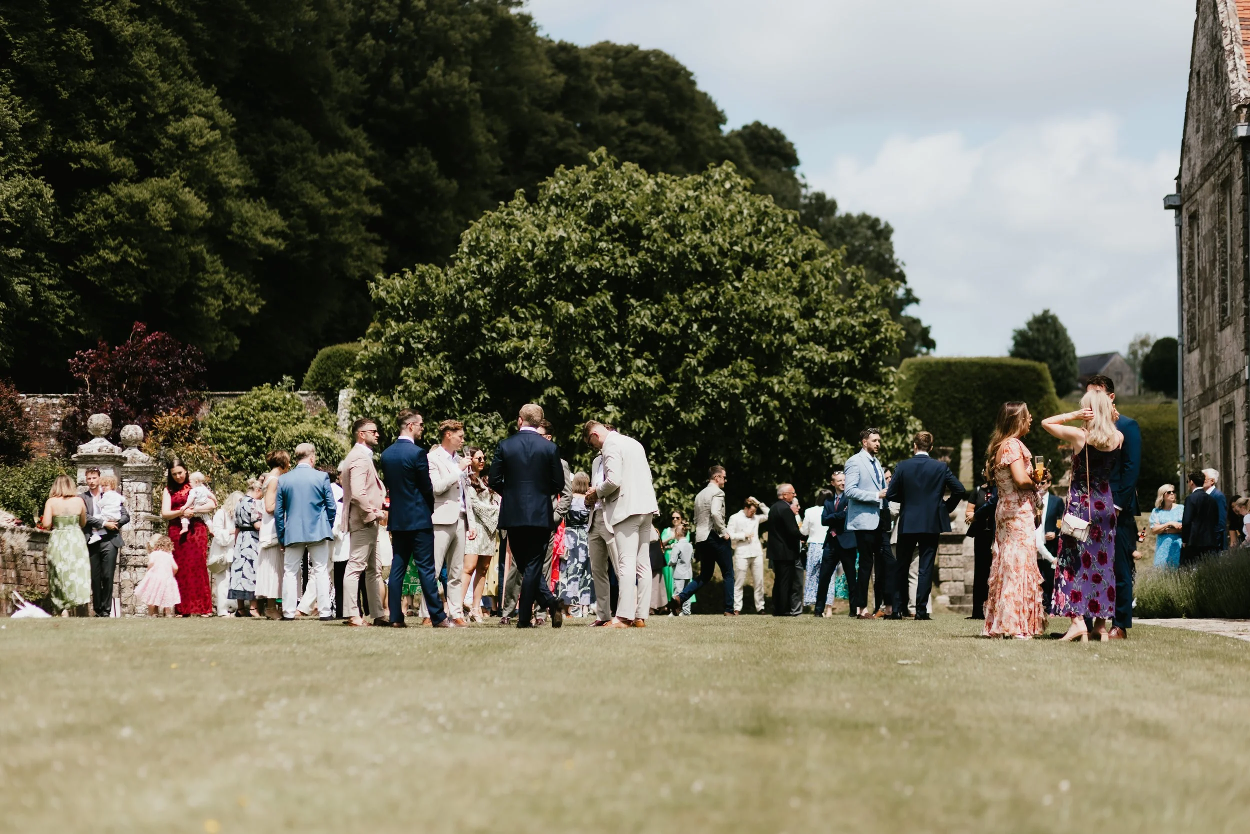 A group of people dressed in formal attire gathered outdoors on a grassy area at Hatch House, with trees and Hatch House in the background.