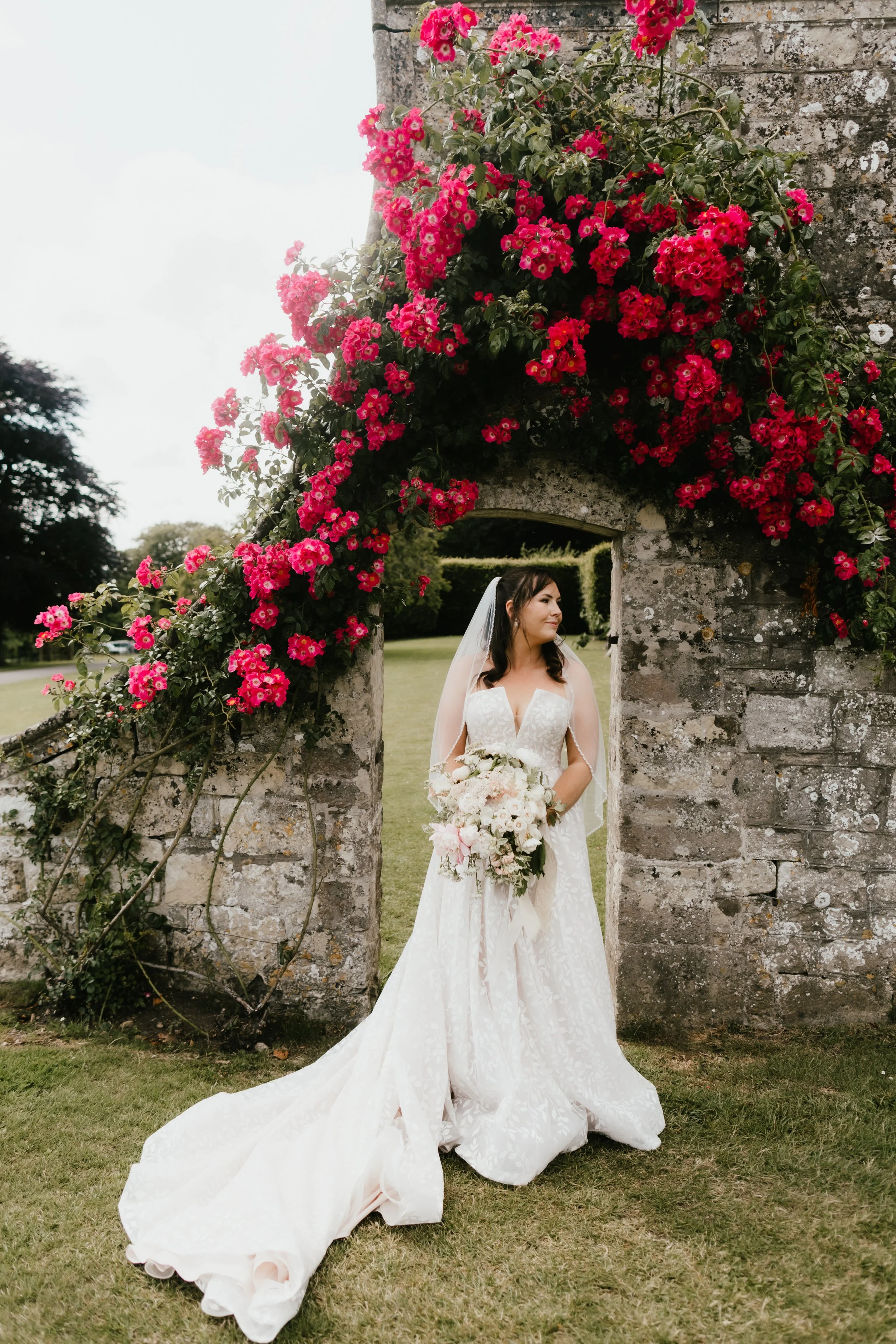 Bride in a white wedding gown holding a bouquet, standing under a stone archway at Hatch House covered with vibrant pink flowers, on a lush green lawn.