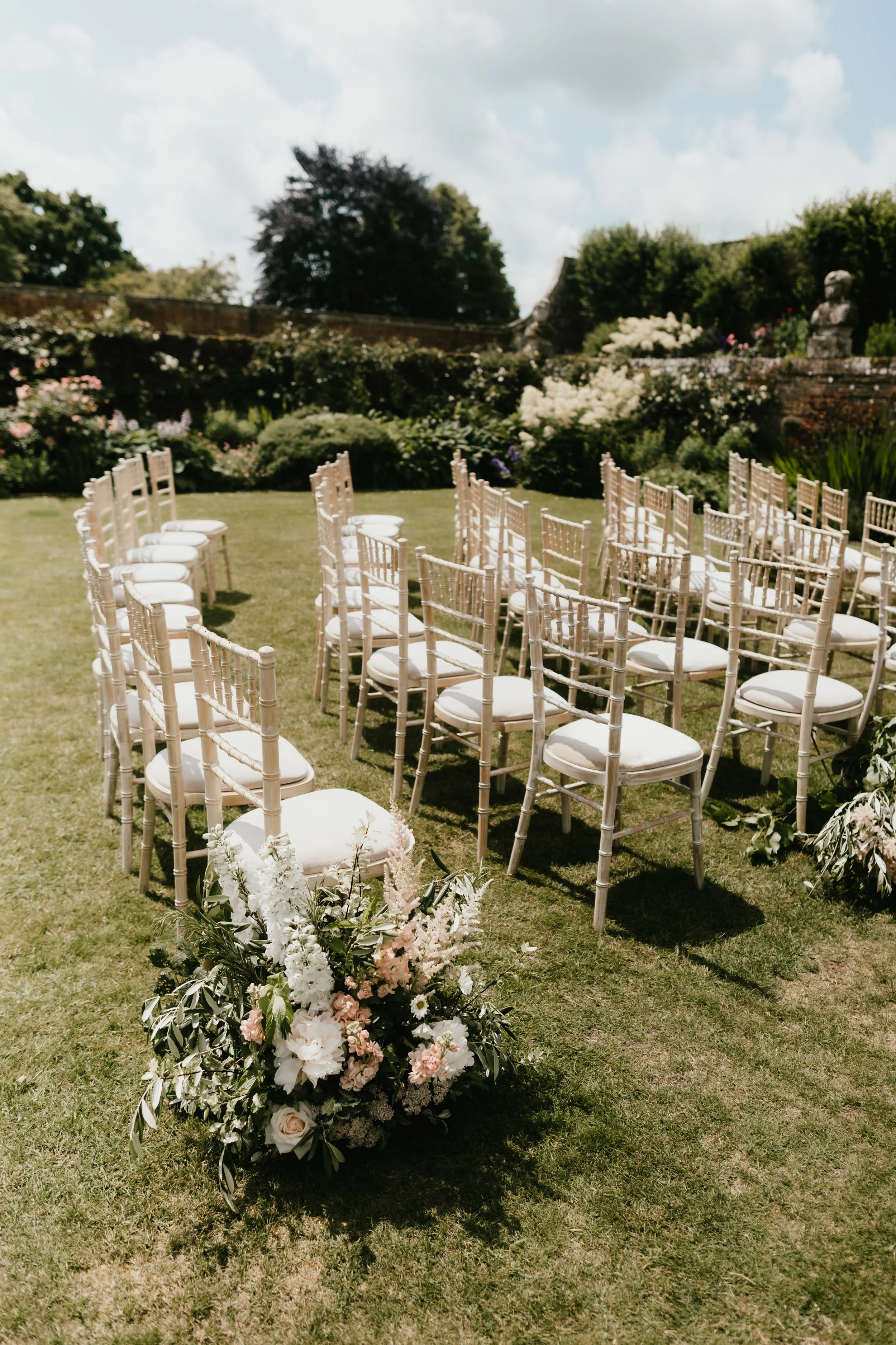 Outdoor wedding setup with white chairs arranged in an aisle, decorated with a floral bouquet at the front, on a grassy area at Hatch House with trees and shrubs.