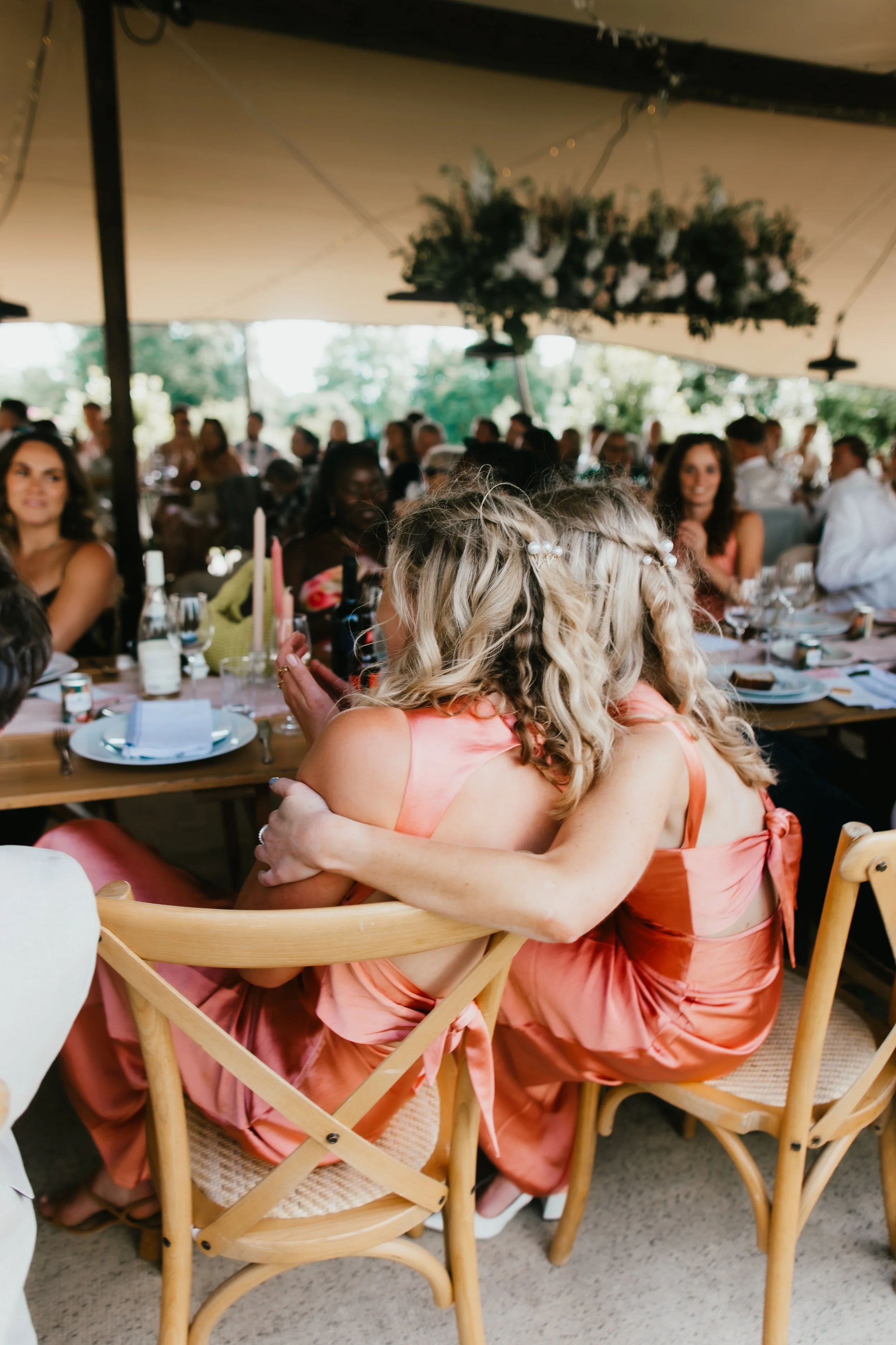Two women with blonde hair, wearing pink dresses, embracing at a festive gathering or celebration, surrounded by seated guests at Pythouse Kitchen Garden.
