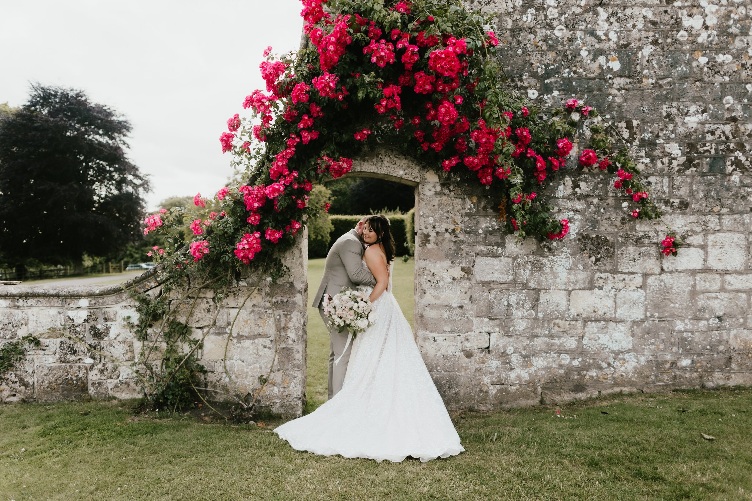 A wedding couple hugging under a stone archway covered with pink roses and greenery, with a grassy area and trees in the background.