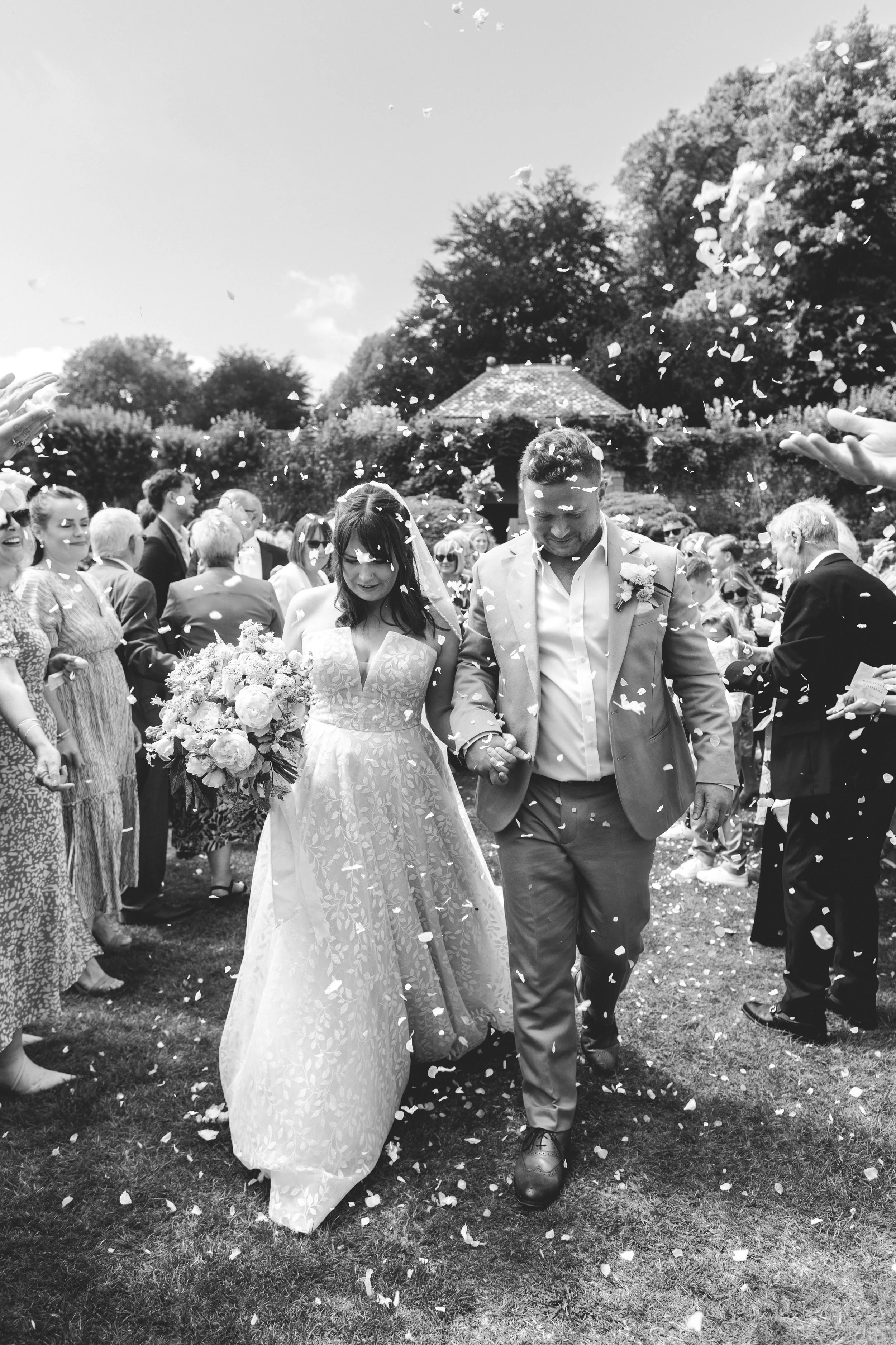 A bride and groom walking hand-in-hand through a crowd of wedding guests, with confetti falling around them in an outdoor setting.