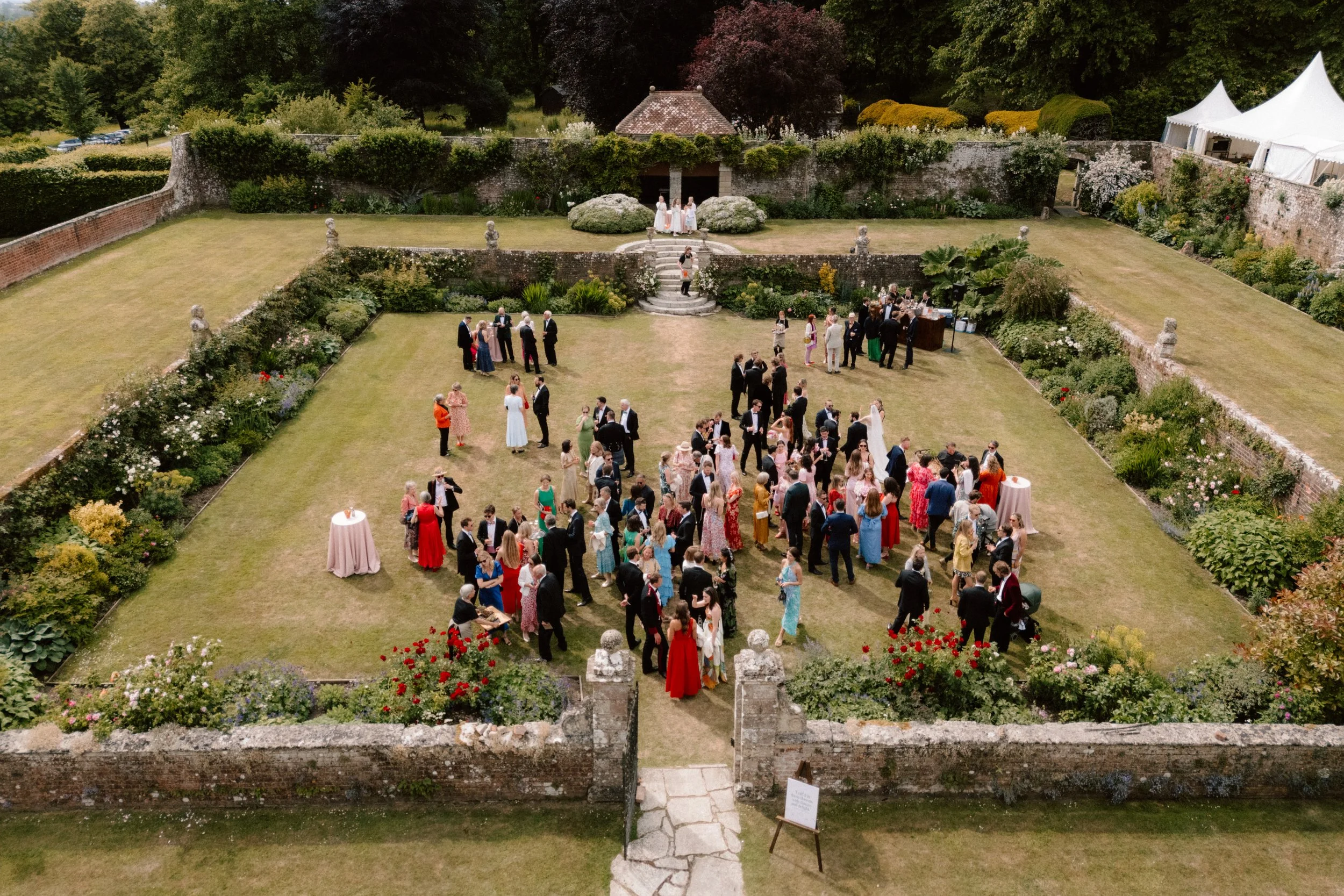 An aerial view of a wedding reception at Hatch House, with guests socialising on the grass, surrounded by blooming flower beds and stone walls.