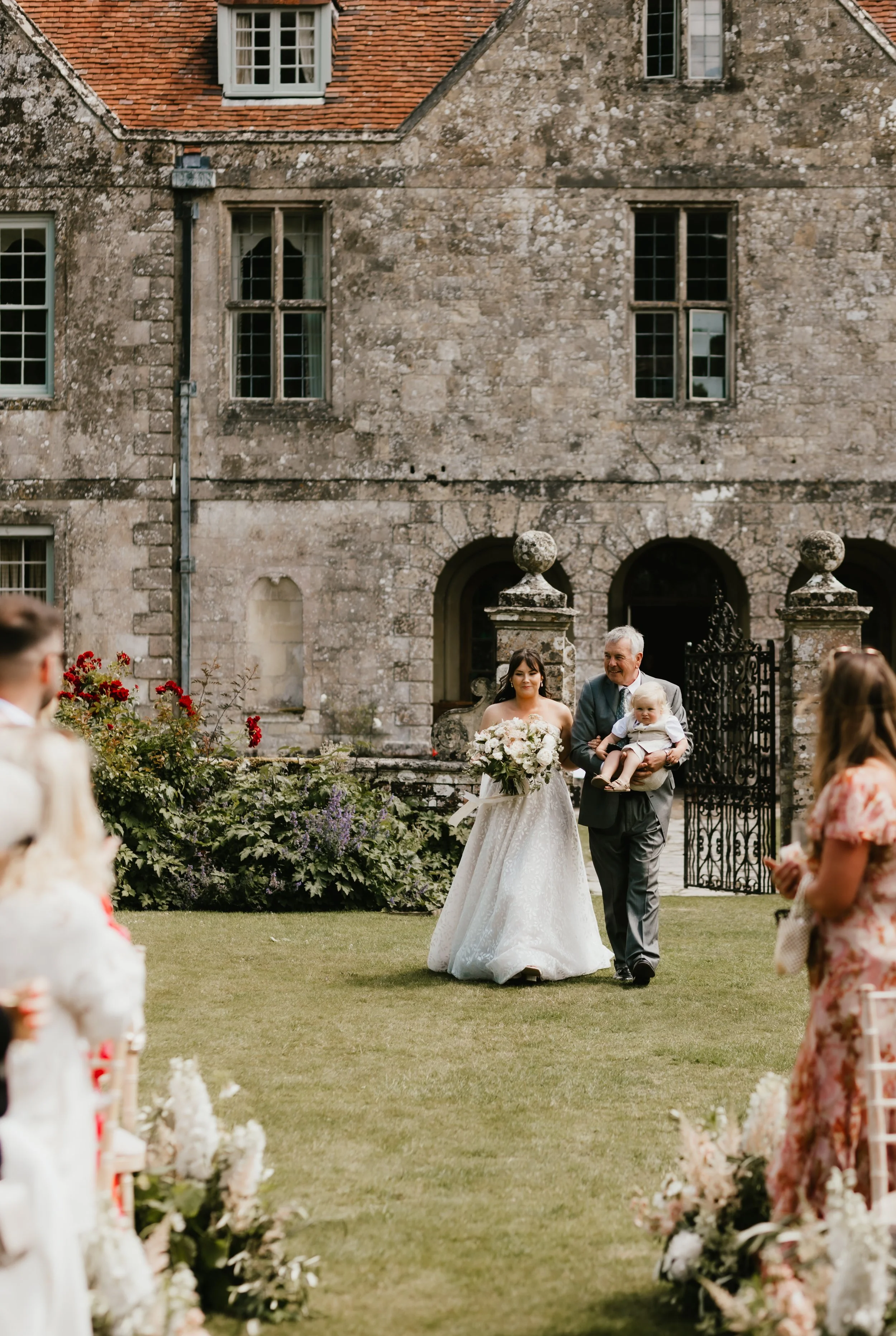 A bride in a white wedding gown holding a bouquet of flowers being escorted by a man with a baby in front of Hatch House during a wedding ceremony.