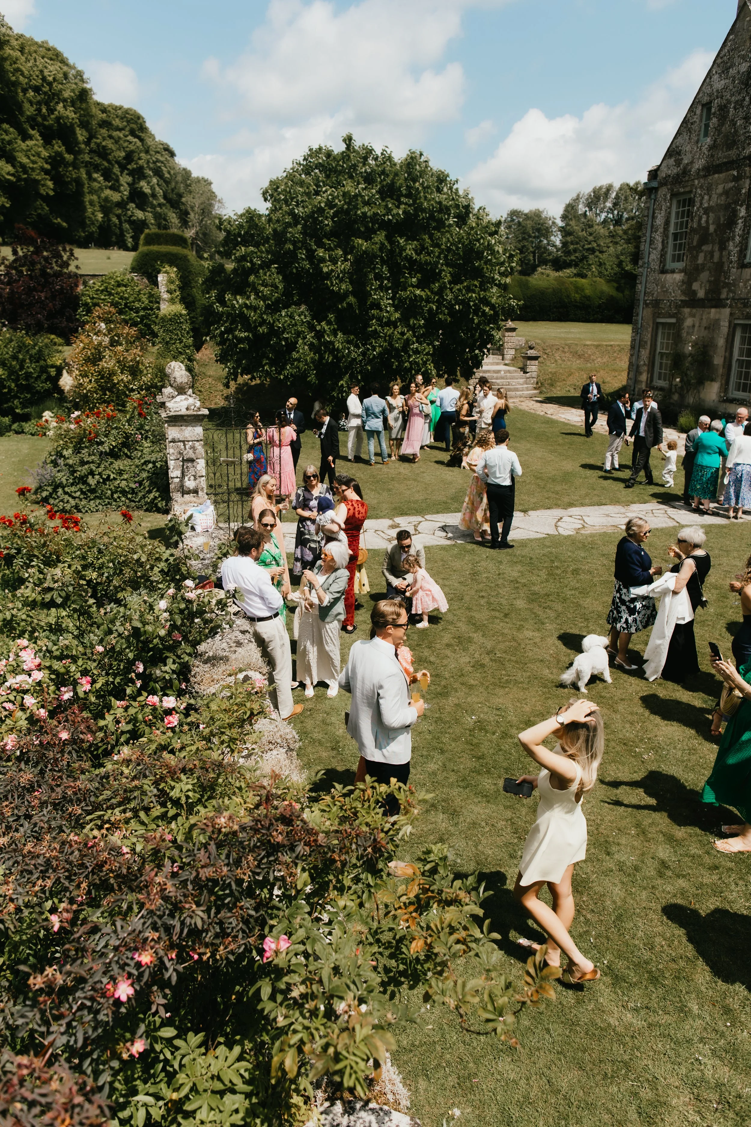 People gathered outdoors on a lush green lawn, some holding drinks, in front of Hatch House during daytime. There are individuals socialising, children, and pets, with a backdrop of trees and a bright blue sky with scattered clouds.