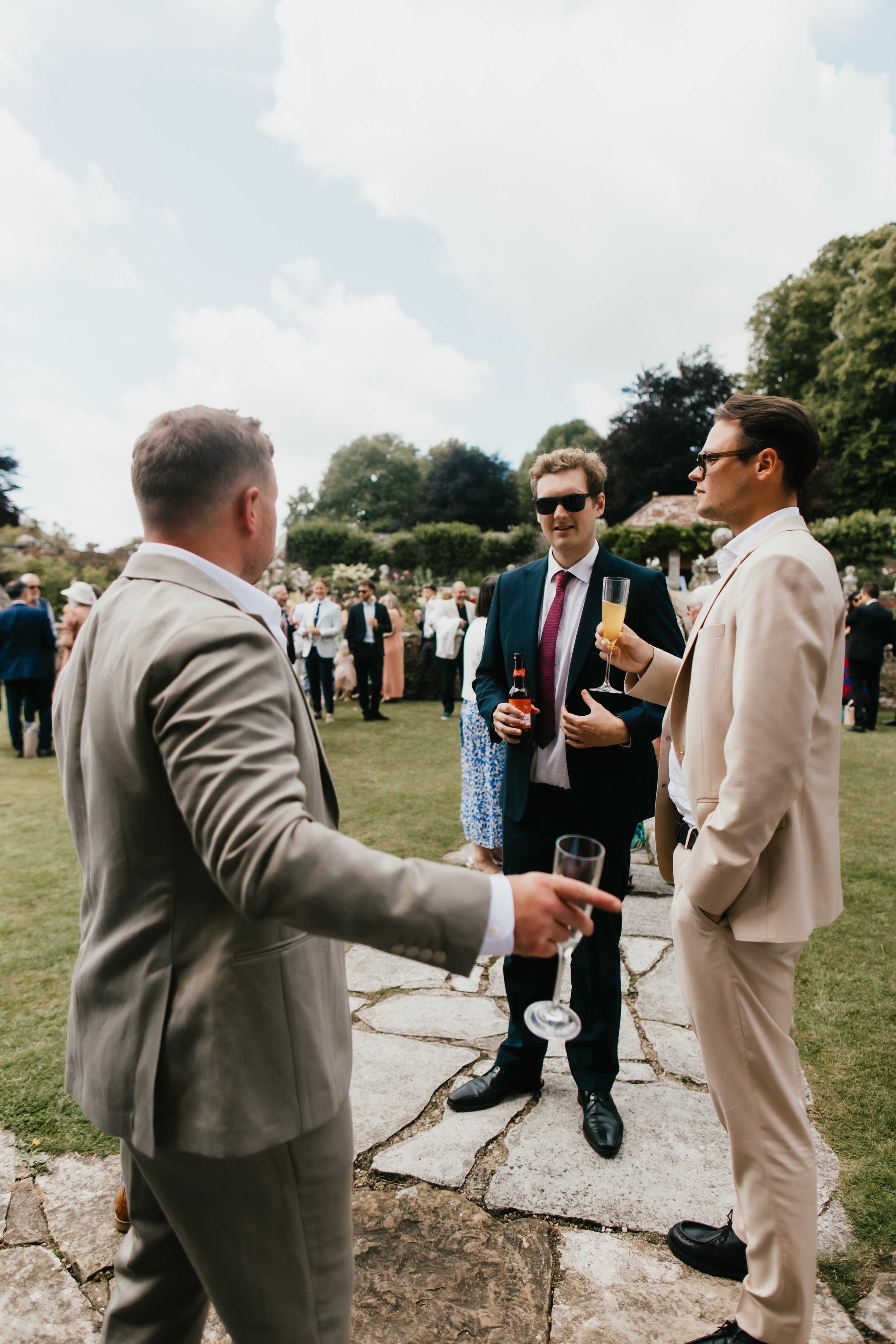 Three men in suits talking and holding drinks at Hatch House with many people in the background under a partly cloudy sky.