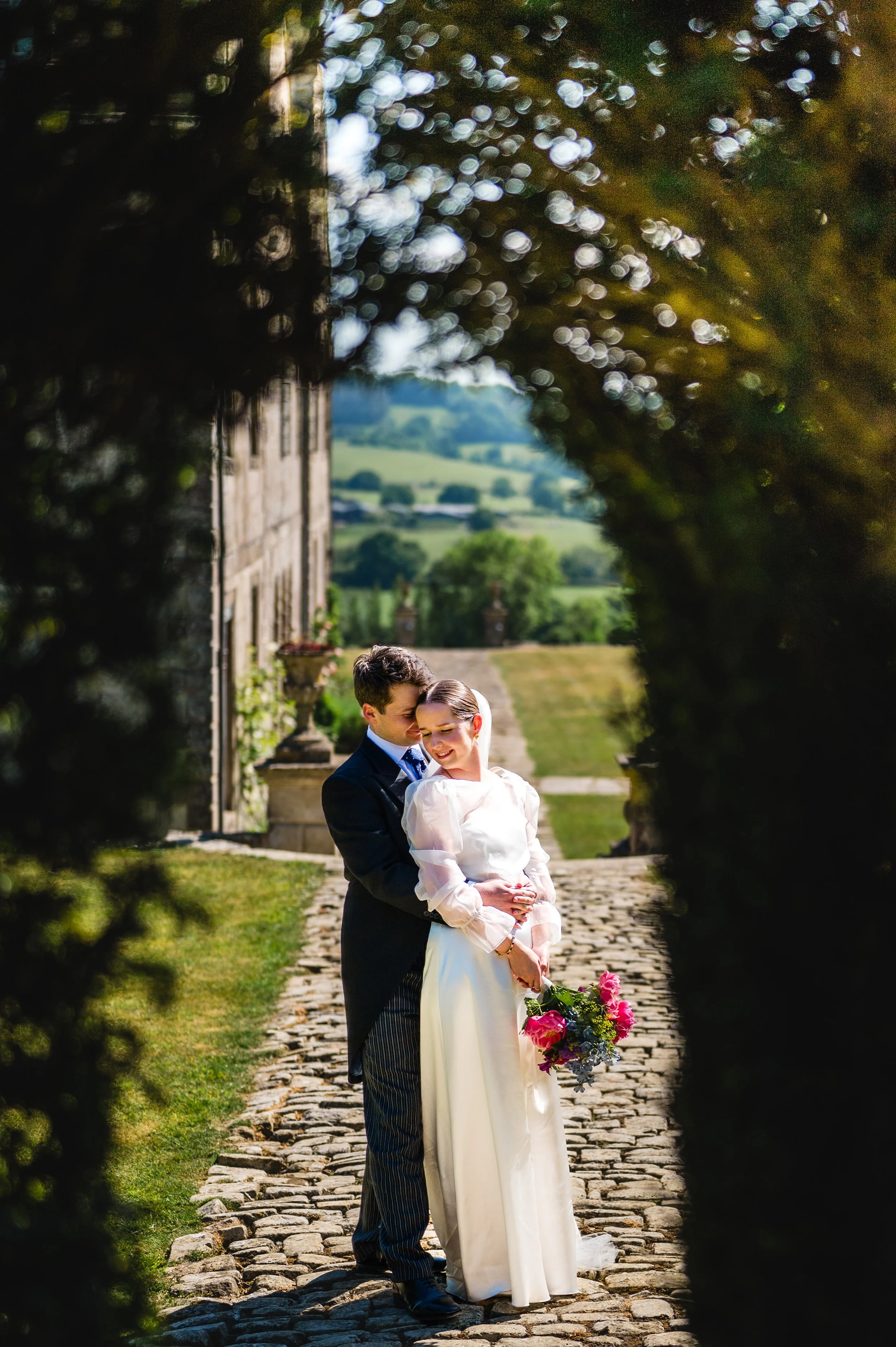 A bride and groom embrace outdoors on a cobblestone path, framed by bushes, with rolling hills in the background on a sunny day.