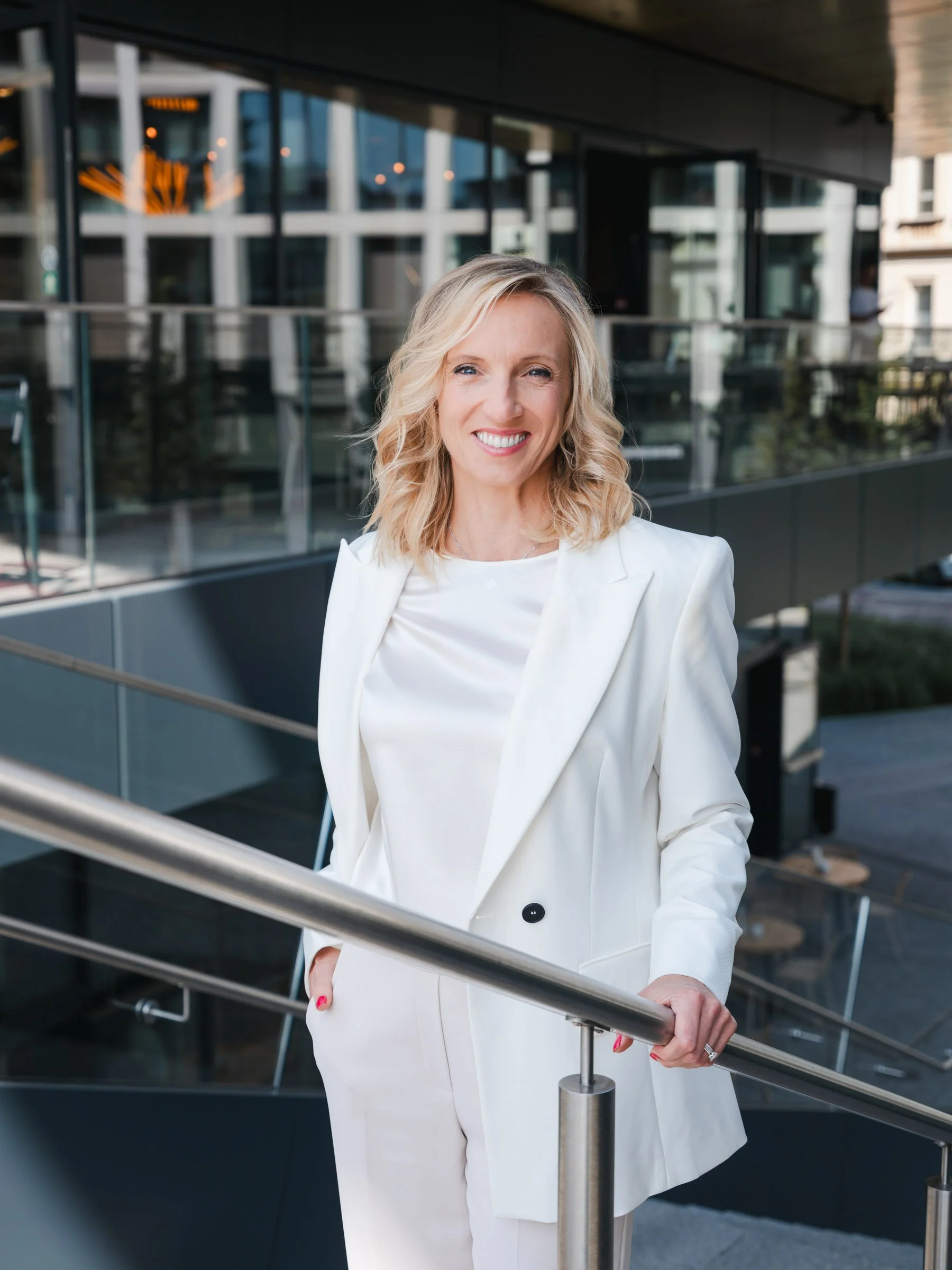A woman with blonde curly hair wearing a white blazer and white pants, smiling and standing outdoors on a staircase with a glass building in the background -business portrait by photographer Dan Hromada from Prague.