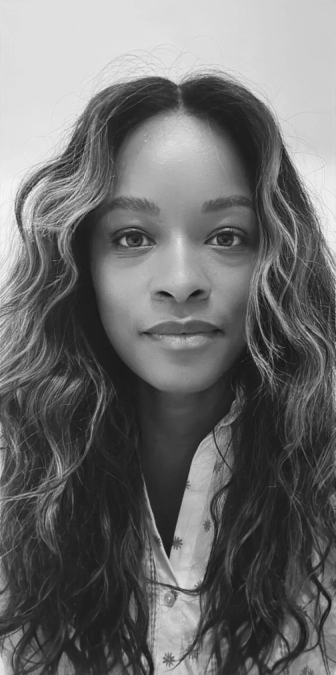Portrait en noir et blanc d'une femme avec de longs cheveux bouclés, portant une chemise à motifs floraux, regardant l'objectif.