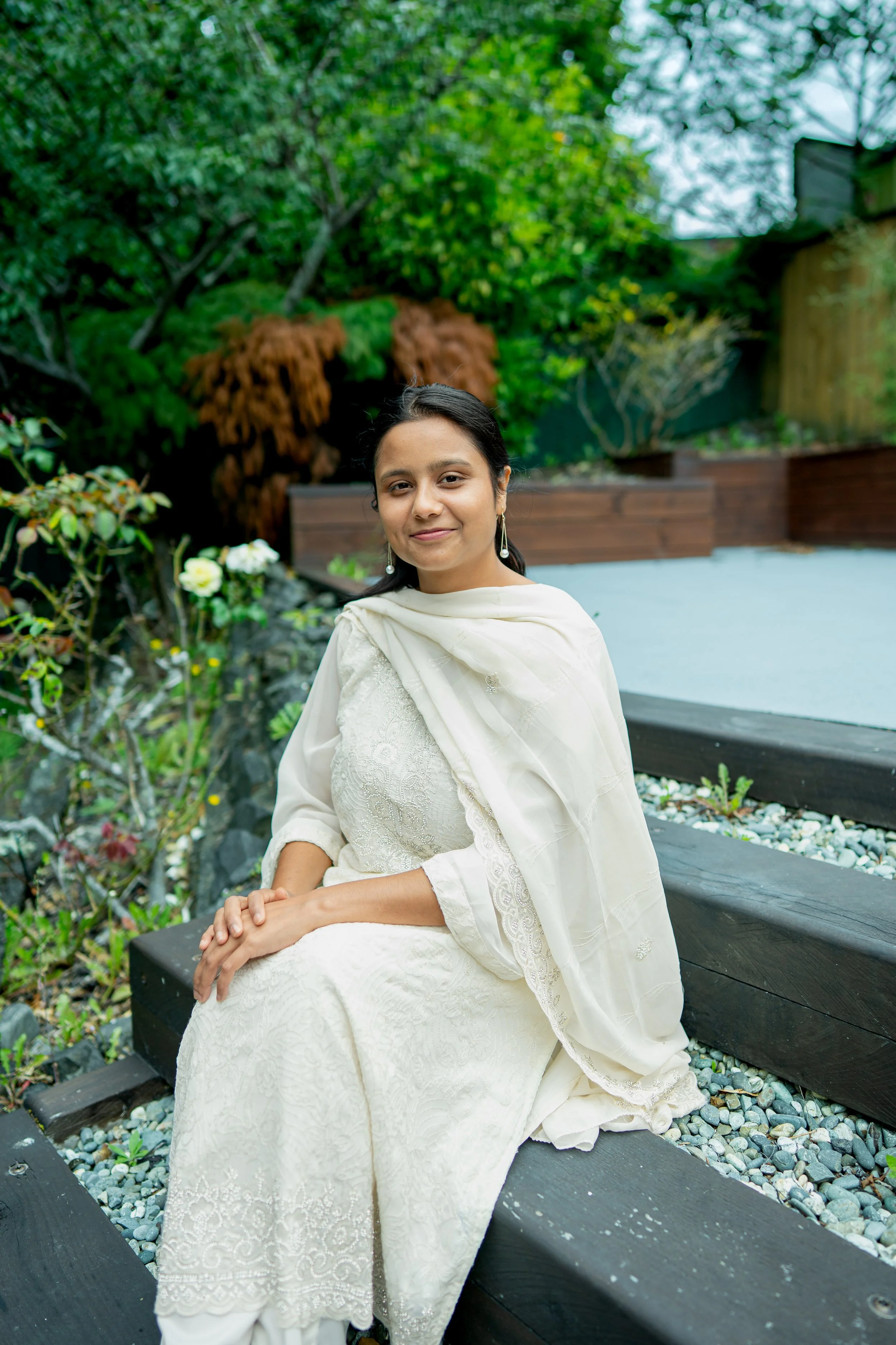 Smiling young woman with black hair and black blazer outdoors with green trees in background