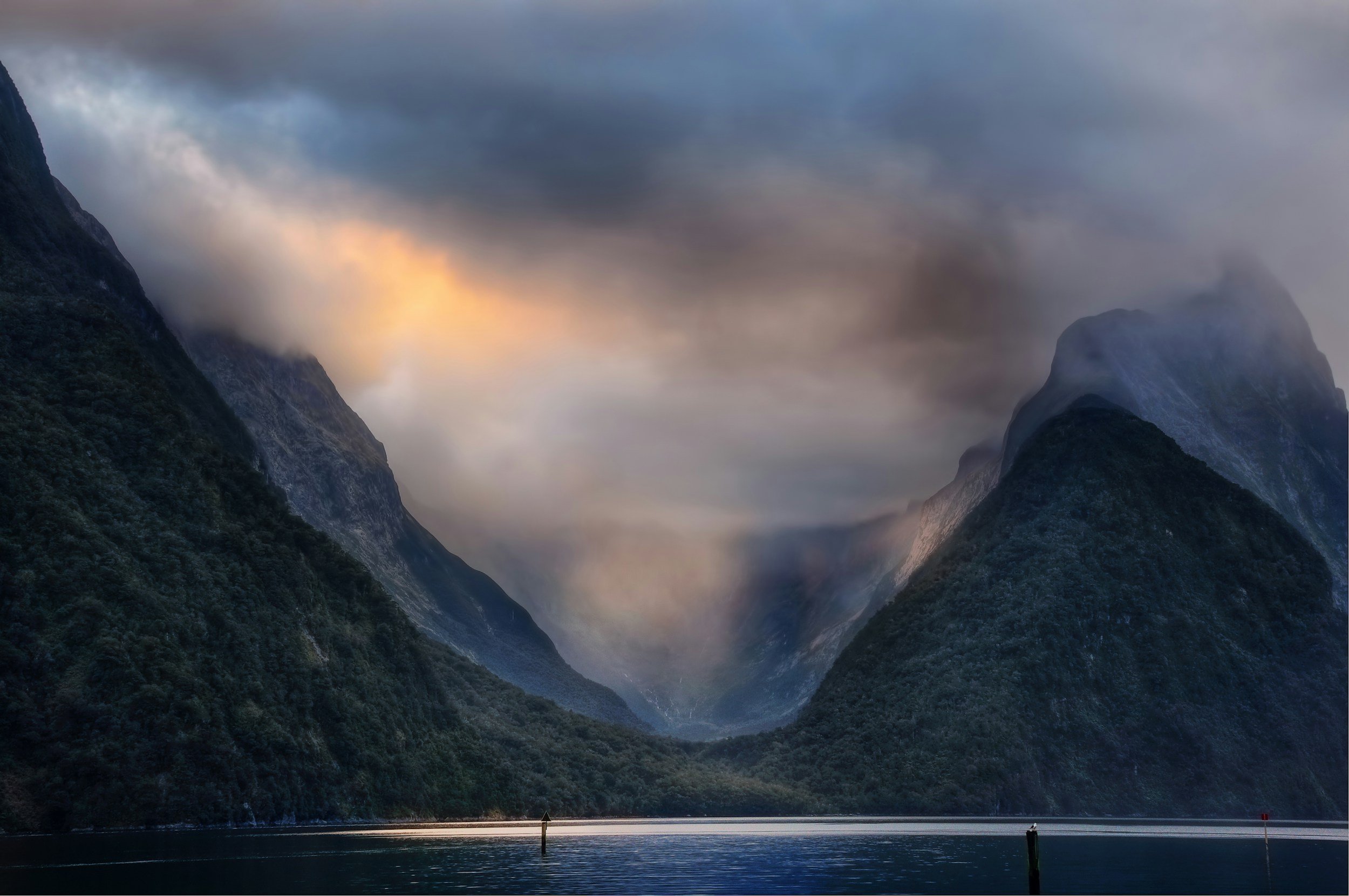Scenic view of a fjord with tall, steep mountains on both sides, partially covered in clouds and mist, reflecting in calm water, with a sky overhead that is partly cloudy with a soft glow.