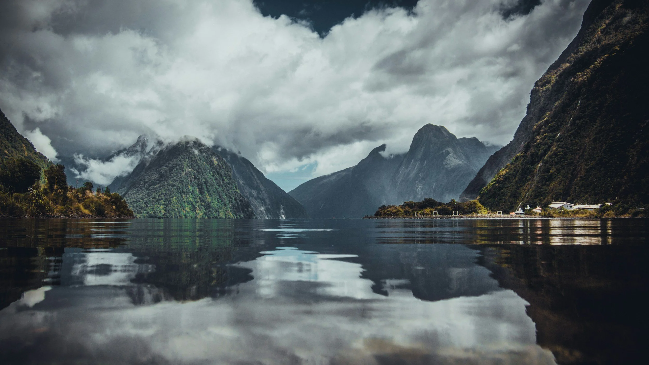 A calm body of water with reflections of green mountains and cloudy sky, with a house and structures on the shoreline to the right.
