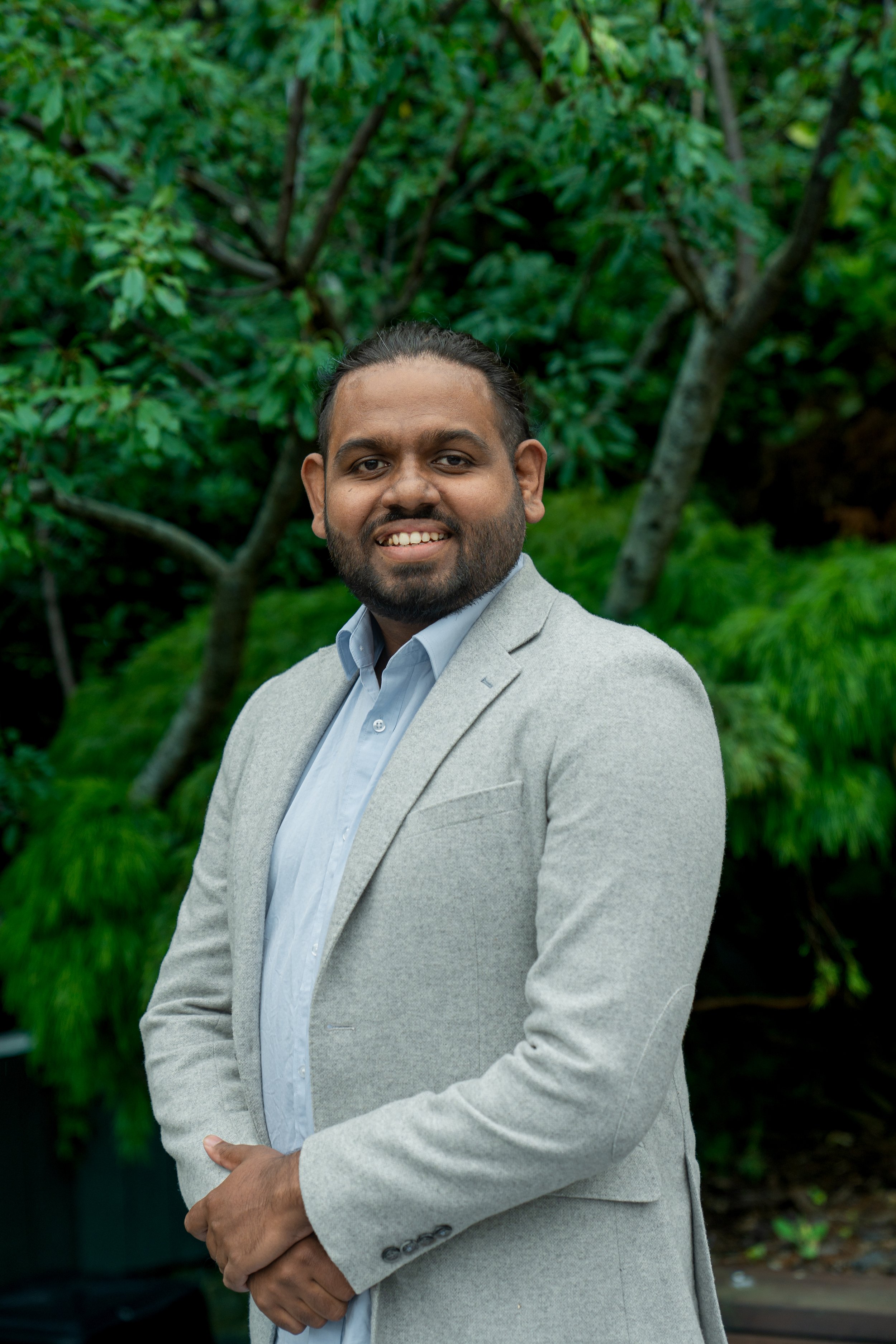 Man with dark hair and a beard, wearing a navy coat, standing outdoors near a body of water and mountains on a cloudy day.
