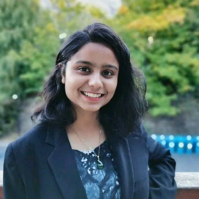Smiling young woman with black hair and black blazer outdoors with green trees in background