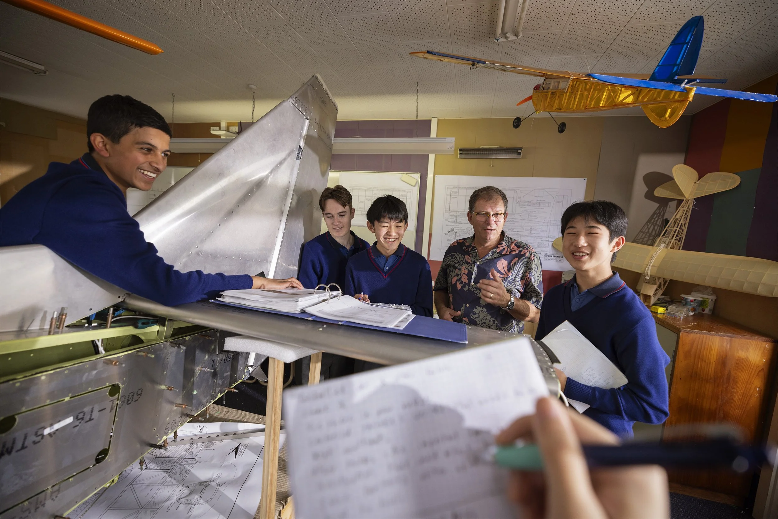 Group of students and an instructor in a classroom with a large model airplane, blueprints, and a yellow remote-controlled airplane hanging from the ceiling, discussing engineering designs.