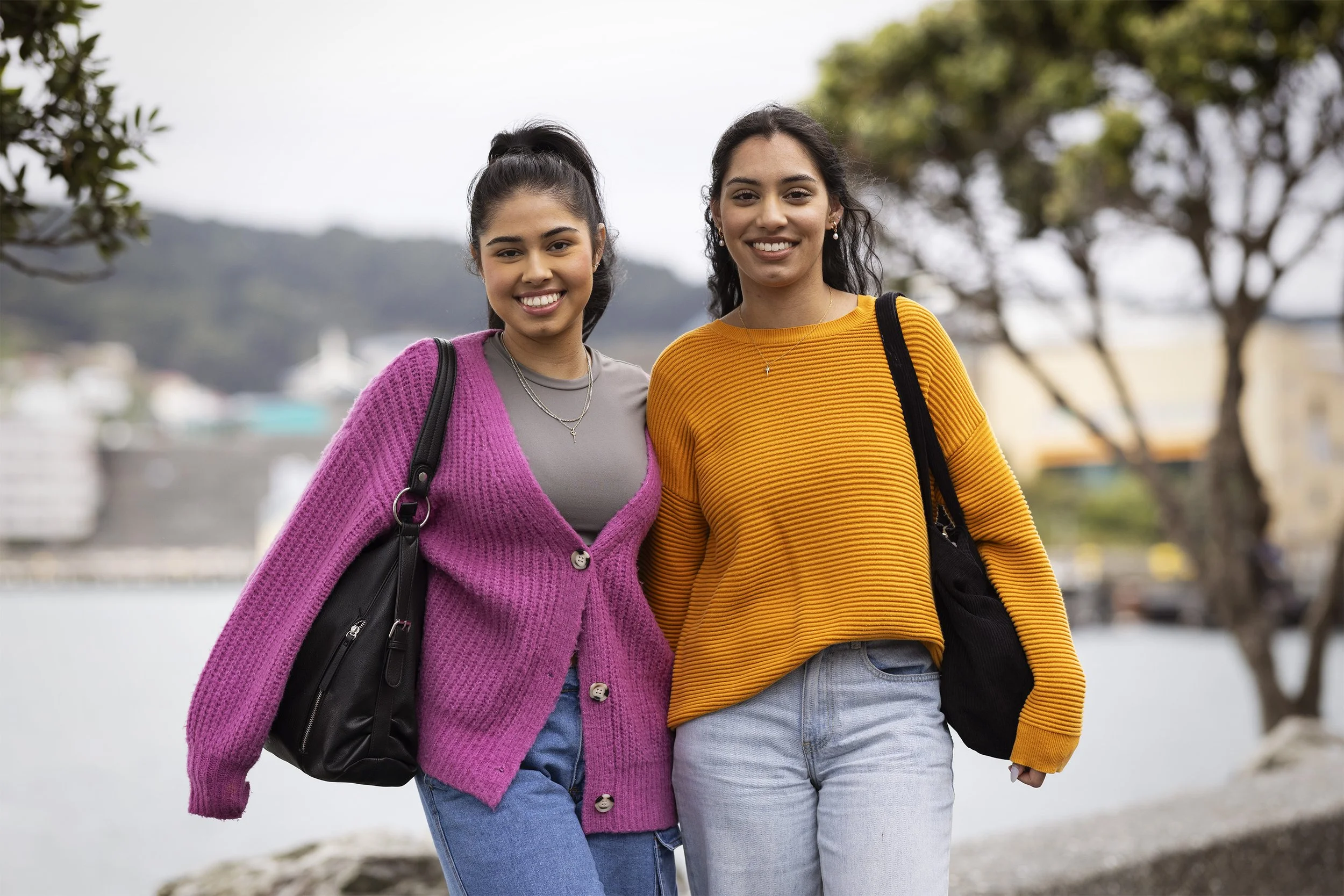 Two young women standing outdoors by a body of water, smiling, with trees and hills in the background. One wears a pink cardigan, the other a yellow sweater, and both carry black bags.