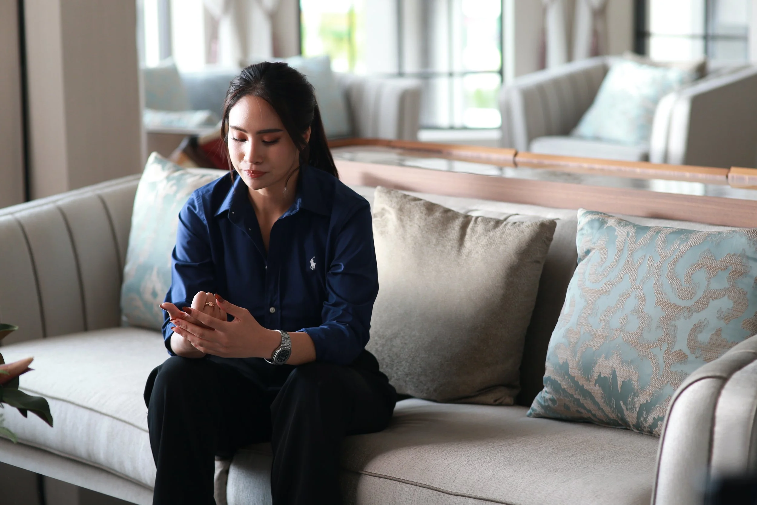 A woman with dark hair in a ponytail, wearing a blue long-sleeve shirt and black pants, sitting on a beige sofa with light-colored and patterned cushions, looking at her phone in a bright room with large windows.