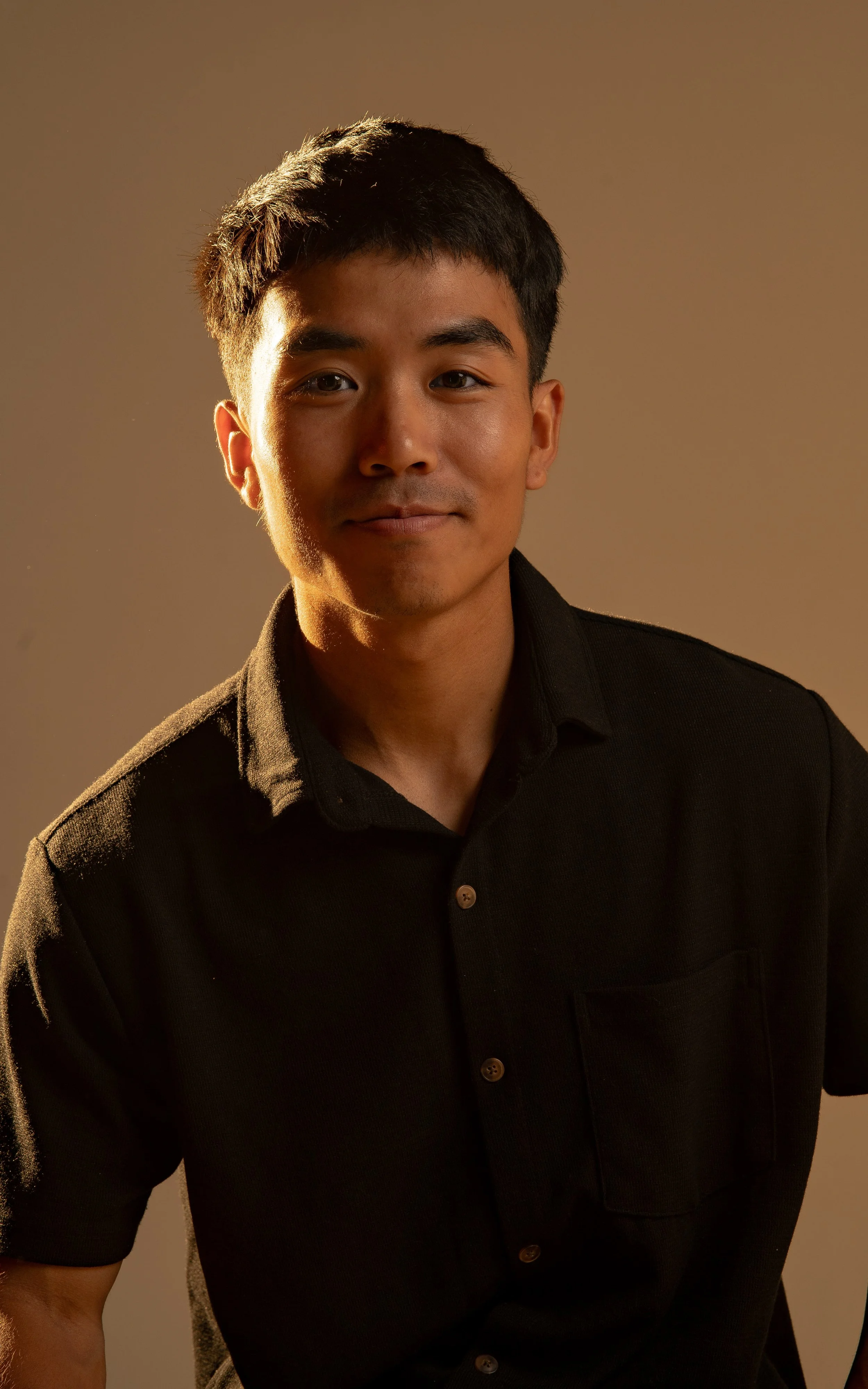 Portrait of a young man with short black hair, wearing a black shirt, looking at the camera with a slight smile, against a neutral background.