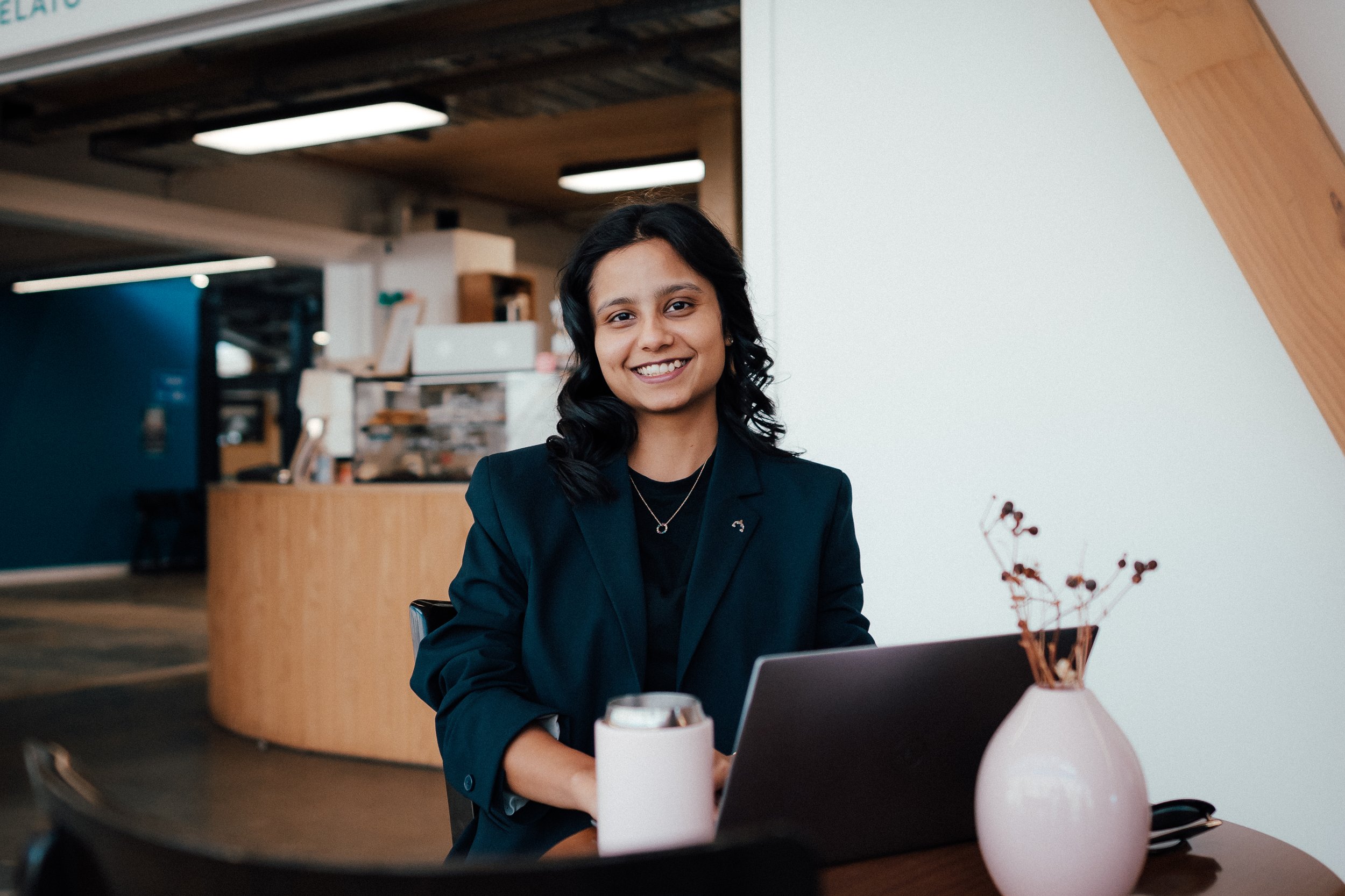 A woman with dark hair, wearing a black blazer and jewelry, smiling while sitting at a table with a laptop and a white vase with dried flowers in a modern office or cafe setting.