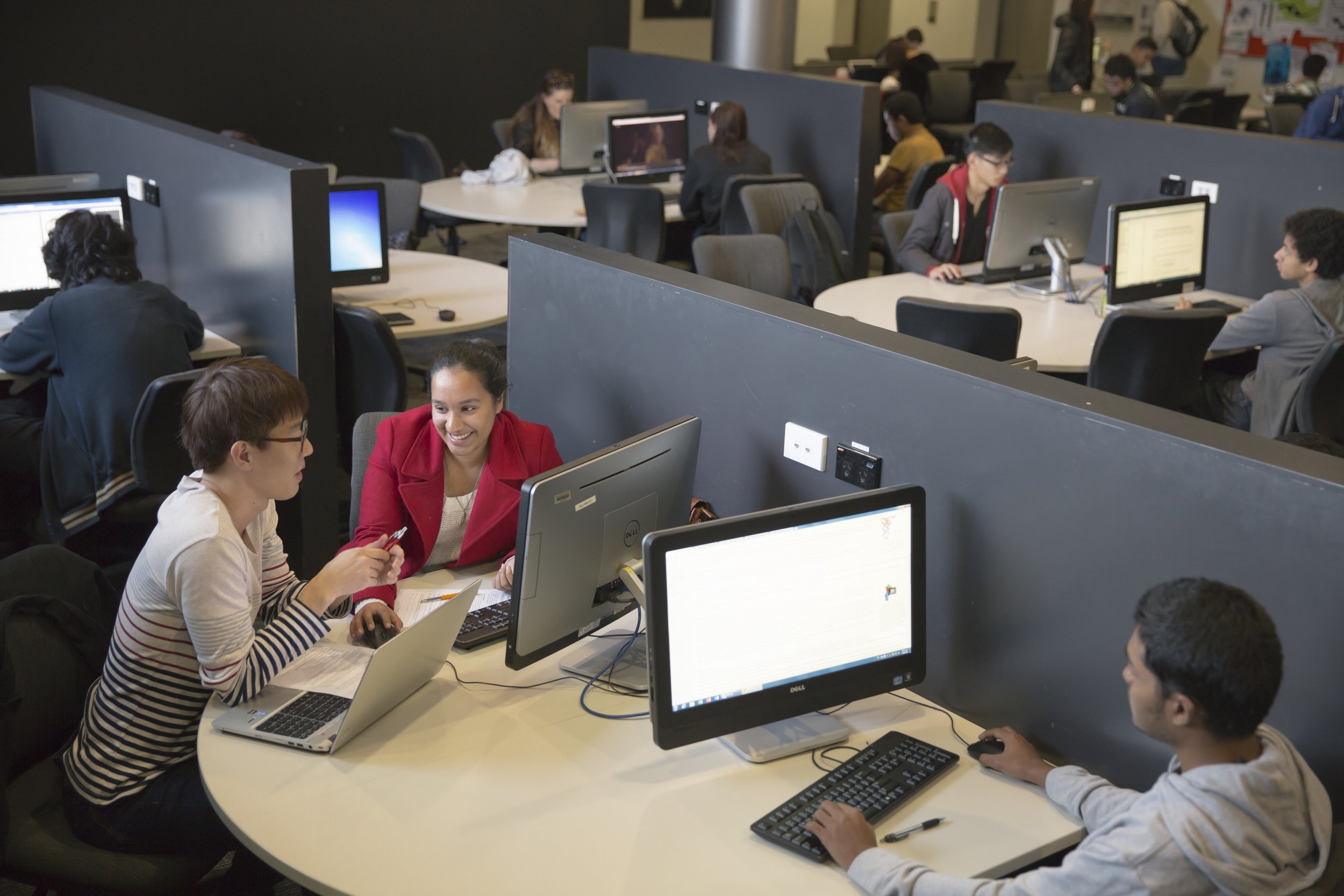 Students working and chatting in a computer lab, with multiple workstations and partitions.