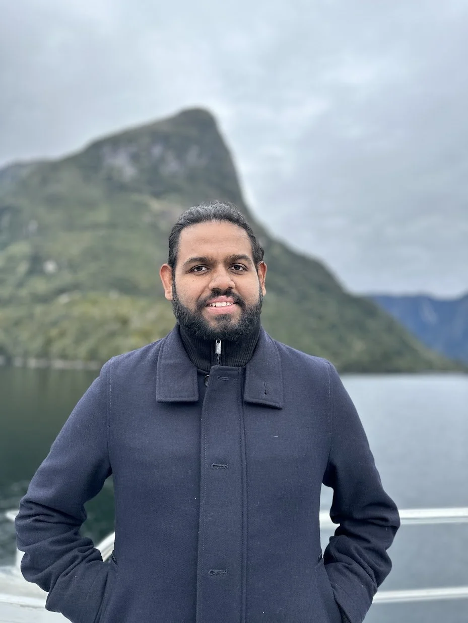 Man with dark hair and a beard, wearing a navy coat, standing outdoors near a body of water and mountains on a cloudy day.