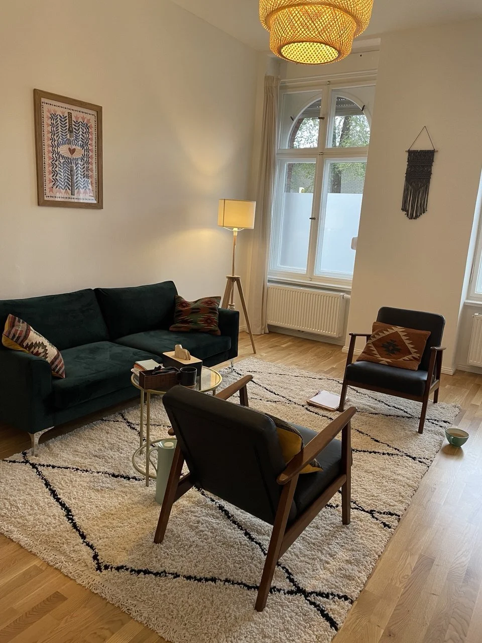 Musiktherapie & Gestalttherapie Praxisraum. Gemütliches Wohnzimmer mit grüner Couch, schwarzen und bunten Kissen, vier Holzsesseln, einem runden Couchtisch, Teppich, Lampe, großen Fenstern und Wanddekoration.