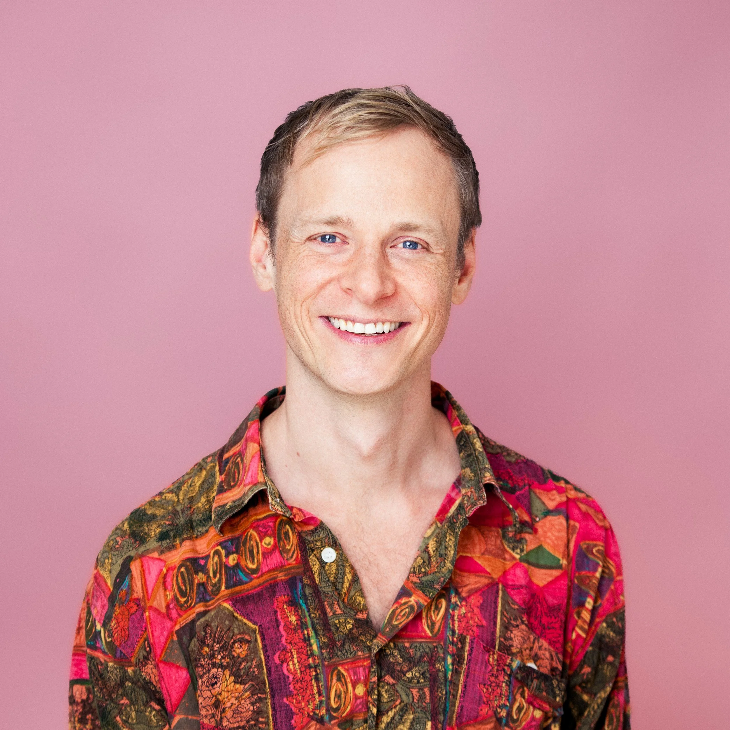 Lächelnder Mann mit blauen Augen und kurzen braunen Haaren trägt ein buntes, Muster-Hemd vor rosa Hintergrund.