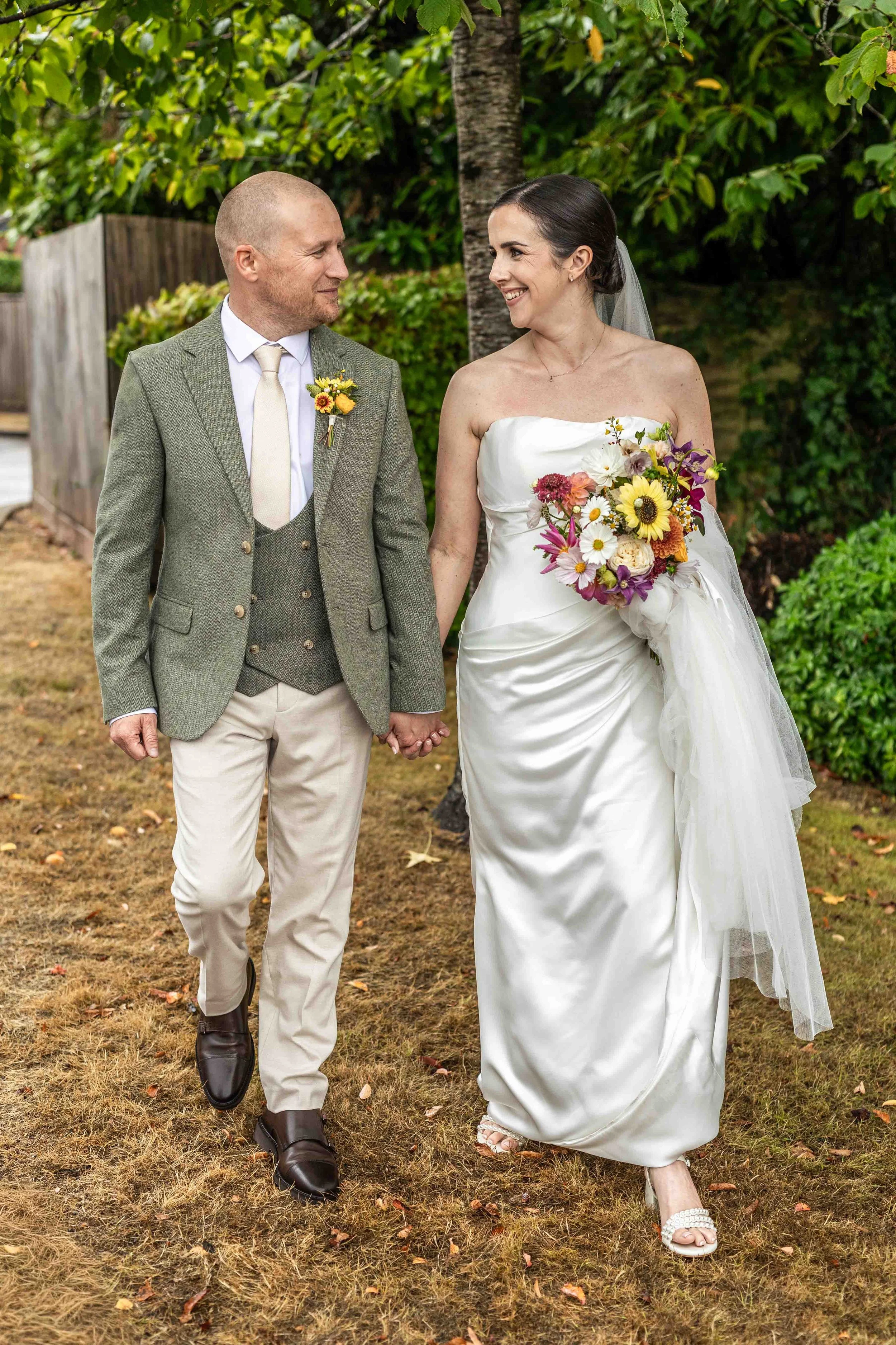 A bride and groom walking hand in hand outdoors during daytime, smiling and looking at each other, surrounded by green foliage and trees, on a grassy ground.