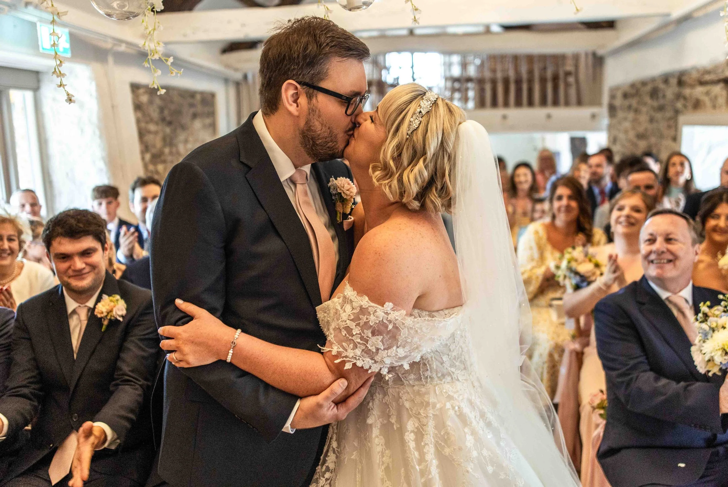 A bride and groom sharing a kiss during their wedding ceremony, with guests seated and smiling in the background.