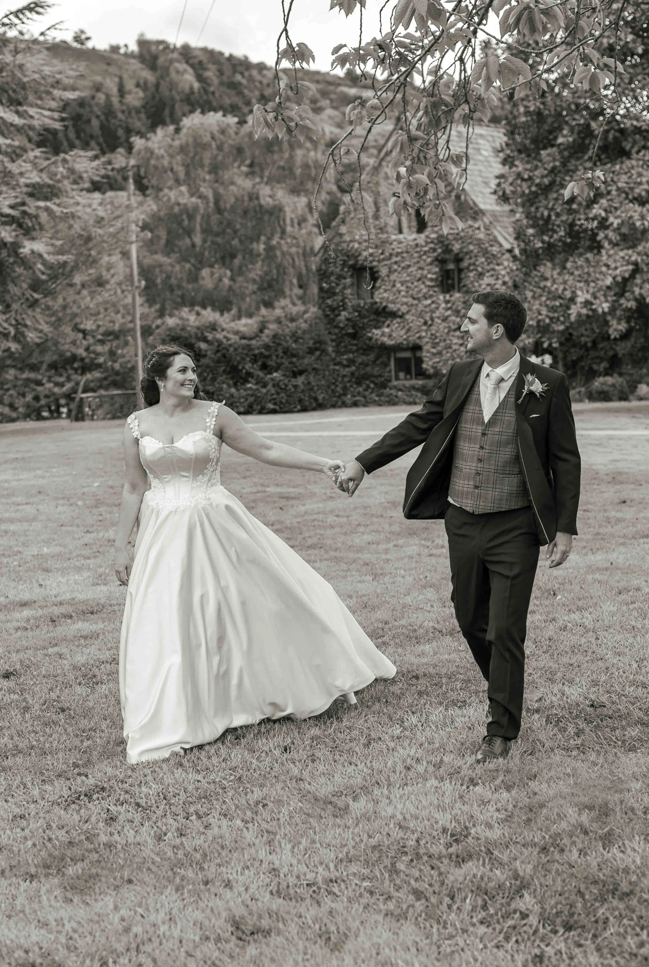 A black and white photo of a bride and groom holding hands and dancing outdoors on a grassy field. The bride is wearing a wedding gown with lace details, and the groom is wearing a suit with a plaid vest and a boutonniere. There are trees and a house