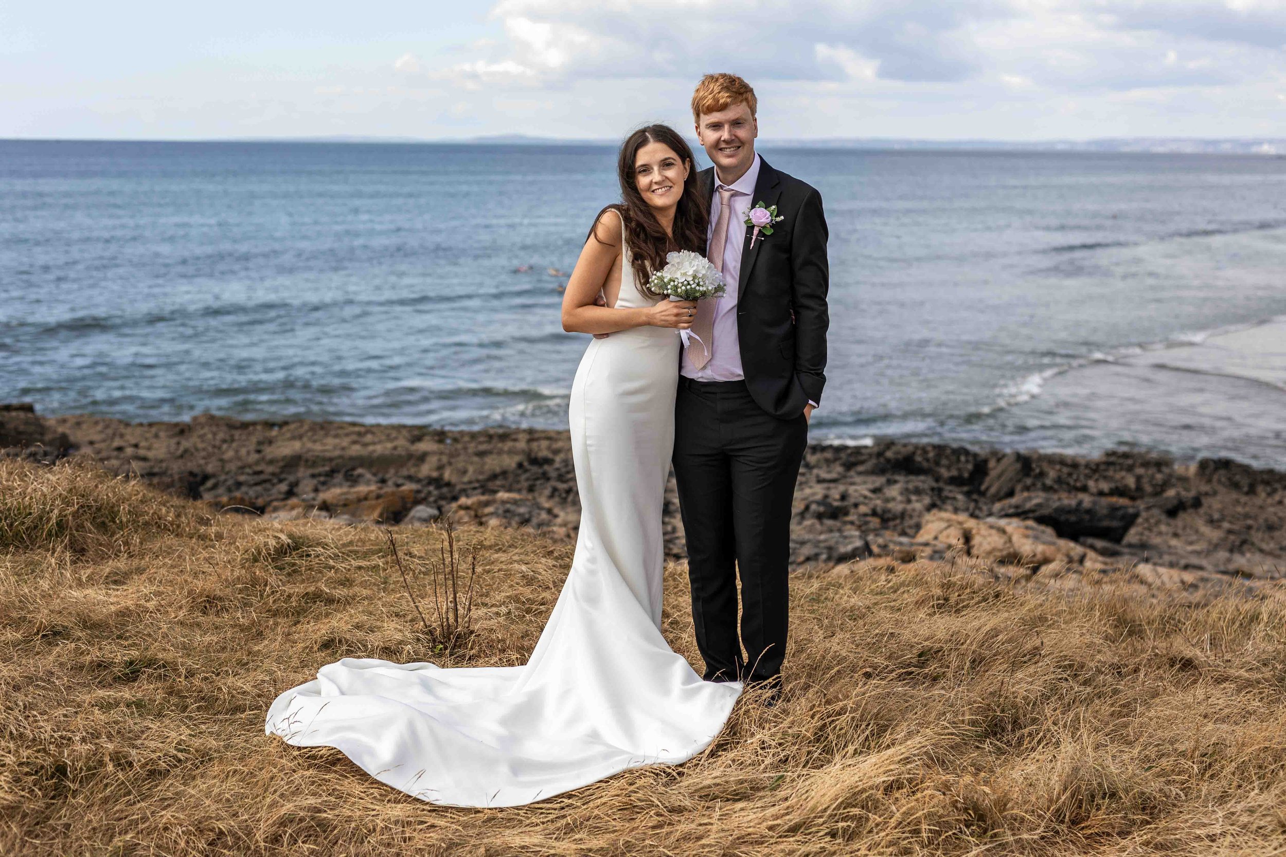 A bride and groom standing on a grassy area near the ocean. The bride is wearing a white wedding gown and holding a bouquet, while the groom is dressed in a suit with a pink tie and a flower boutonniere. They are smiling at the camera with the ocean 