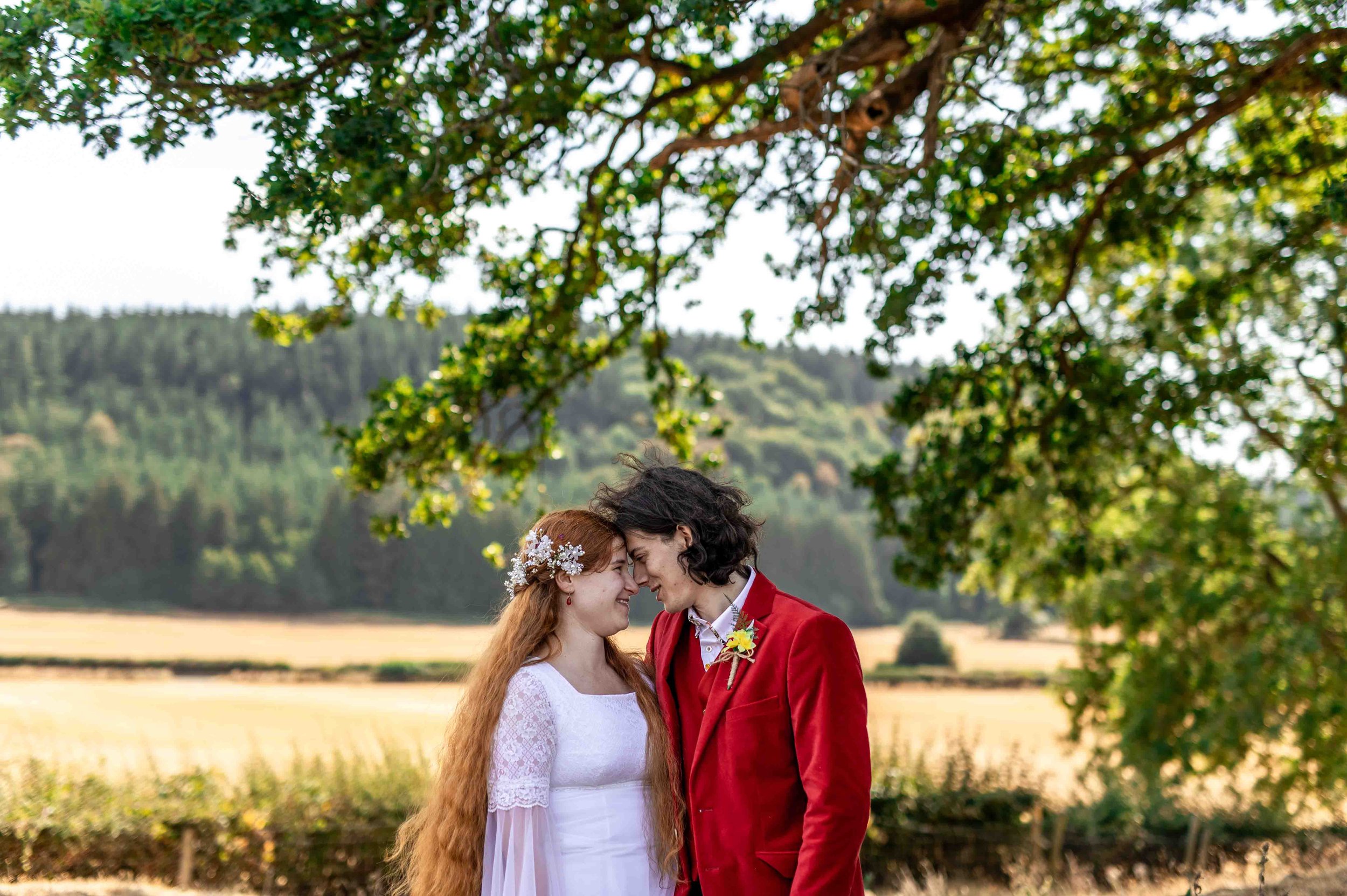 A couple, dressed in wedding attire, standing close with foreheads touching in an outdoor setting with green trees and a field in the background.