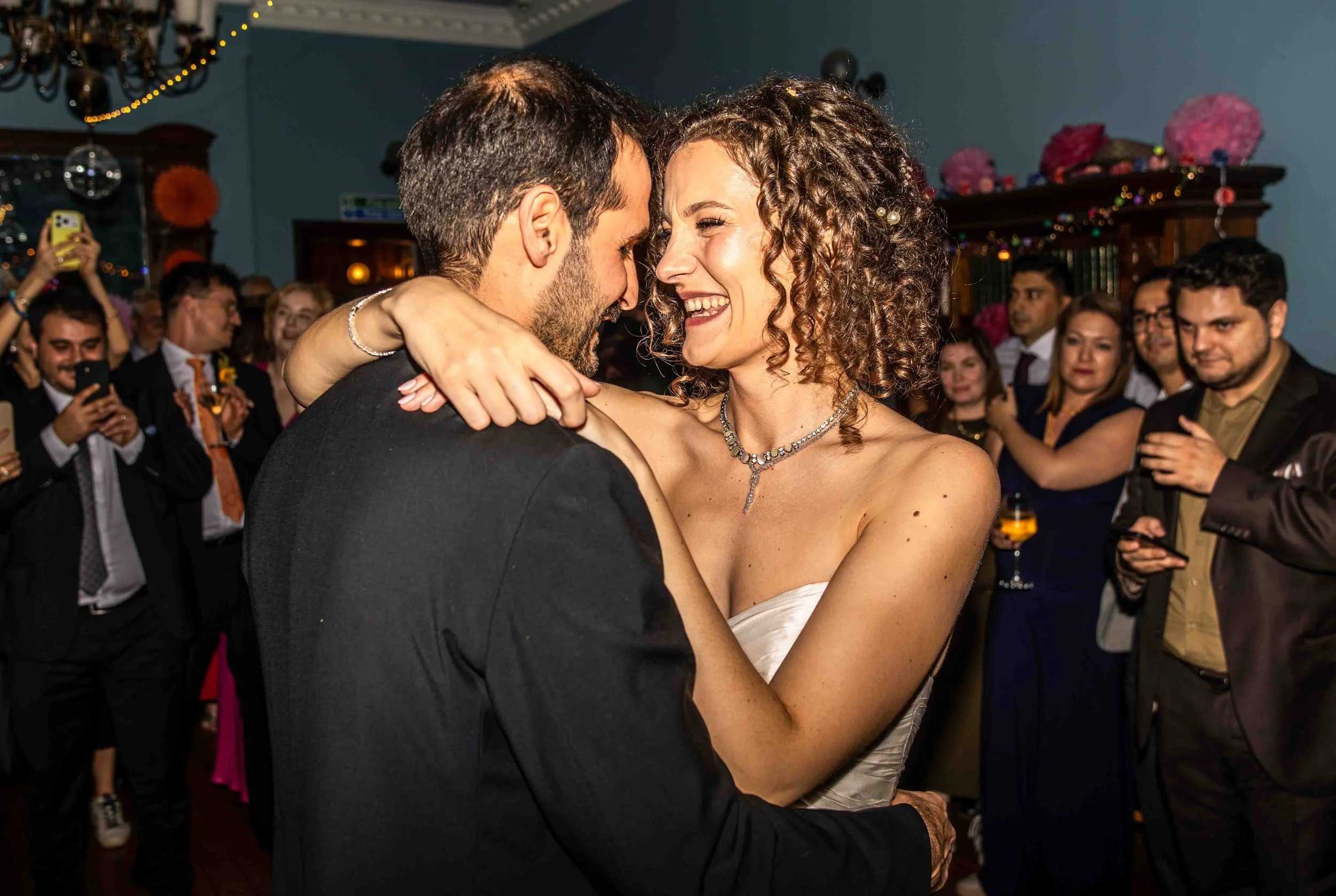 A couple dancing closely at a celebration with many people in the background, some capturing the moment with their phones, in an indoor decorated setting.