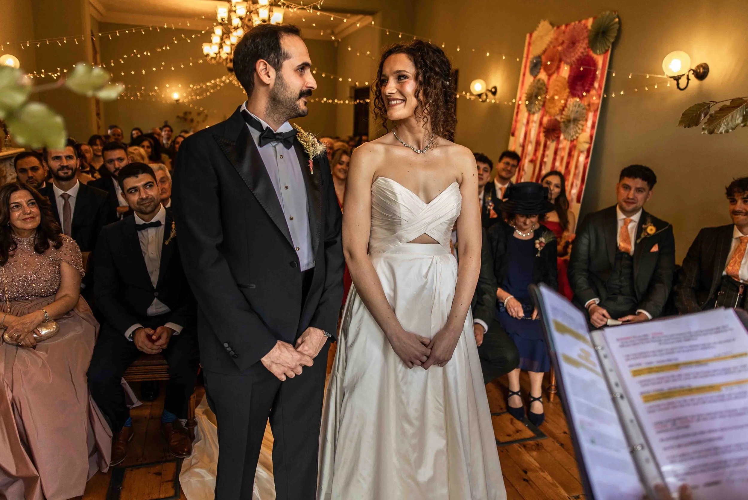 A bride and groom standing together during their wedding ceremony, surrounded by seated guests in a warmly lit room decorated with string lights and artwork.