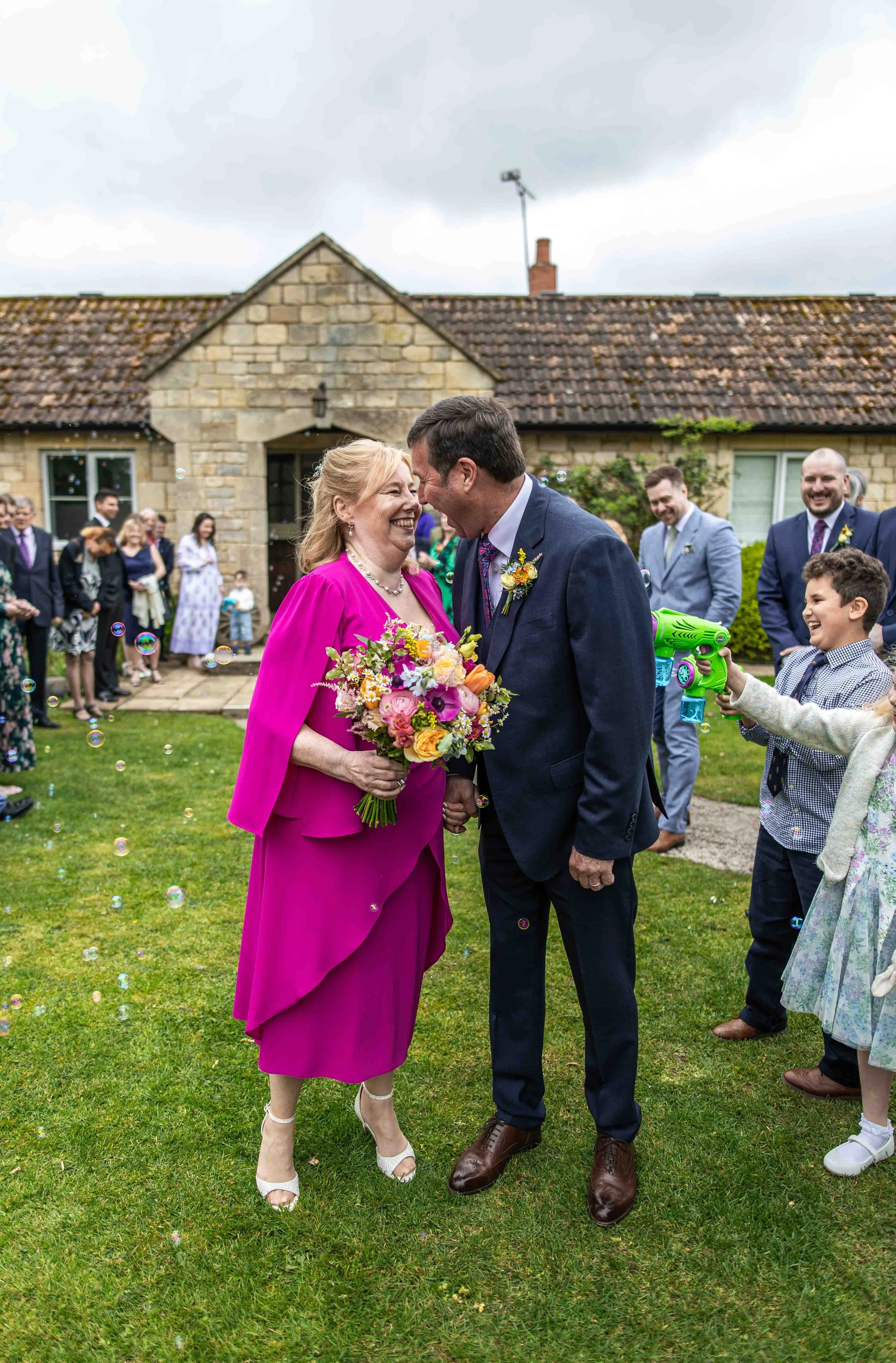 A woman in a pink dress holding a bouquet of flowers is smiling and leaning towards a man in a suit, also smiling, at an outdoor gathering with other people in the background. The setting appears to be a backyard with a stone house and cloudy sky.