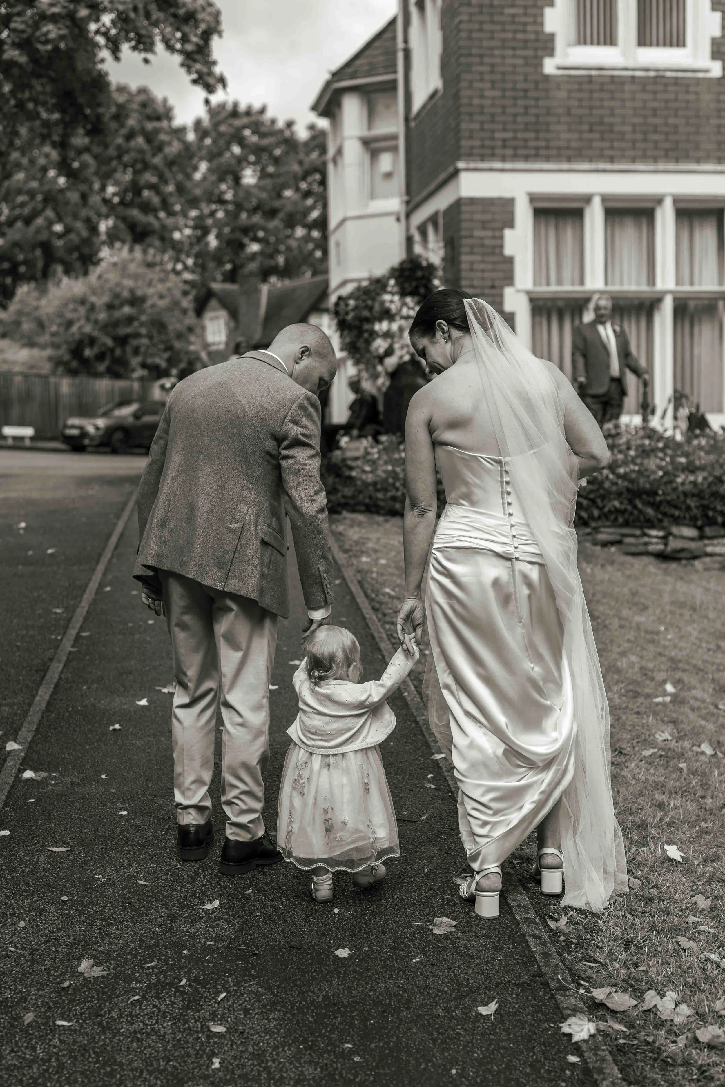 A bride and groom, with a young girl, walk on a sidewalk in a residential area. The couple is dressed in wedding attire, and the girl is in a dress. The photo is in black and white.