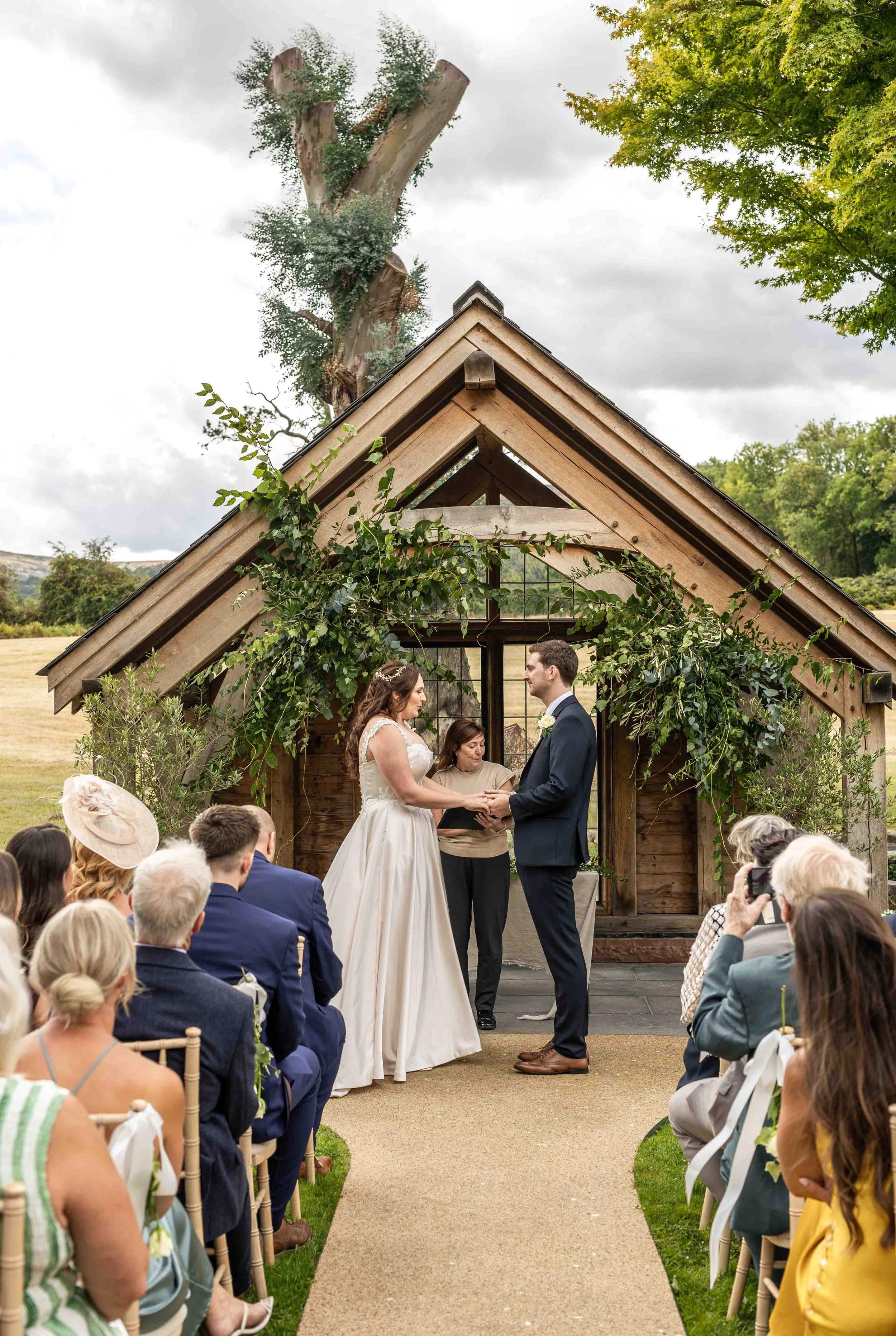 A wedding ceremony taking place outdoors under a wooden structure decorated with greenery. A bride and groom are standing in front of an officiant, exchanging vows, with guests seated on either side.