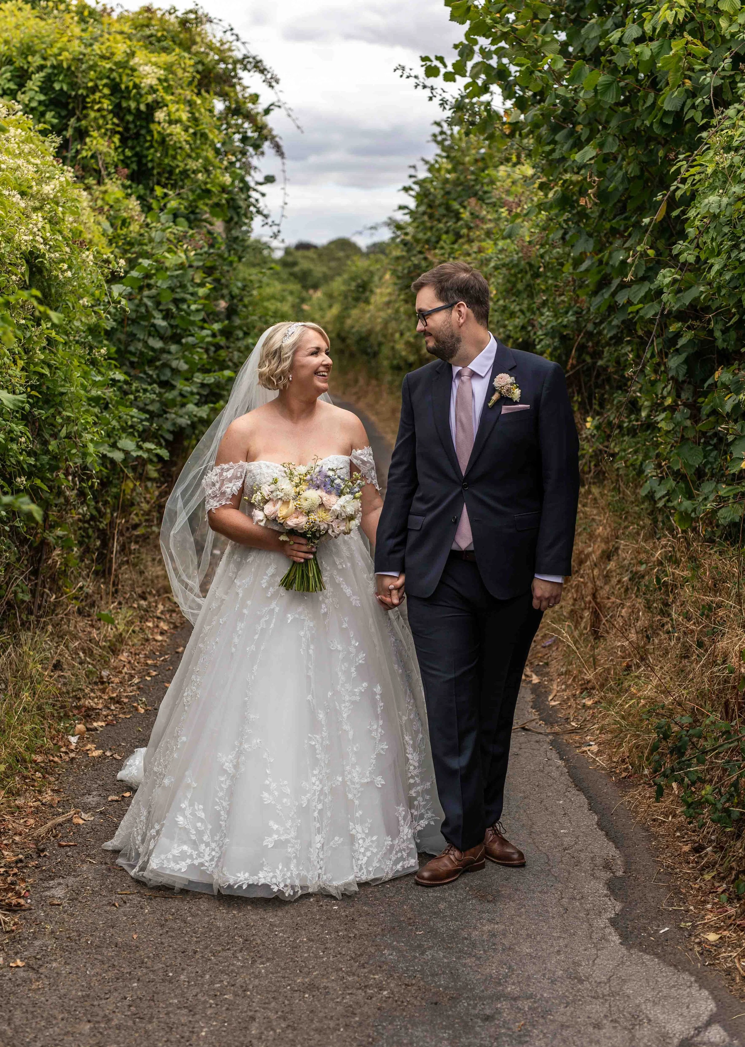 A bride and groom holding hands and smiling at each other while walking on a narrow dirt path surrounded by green bushes during their wedding. The bride is wearing a white lace wedding gown and a veil, and holding a bouquet of flowers. The groom is d
