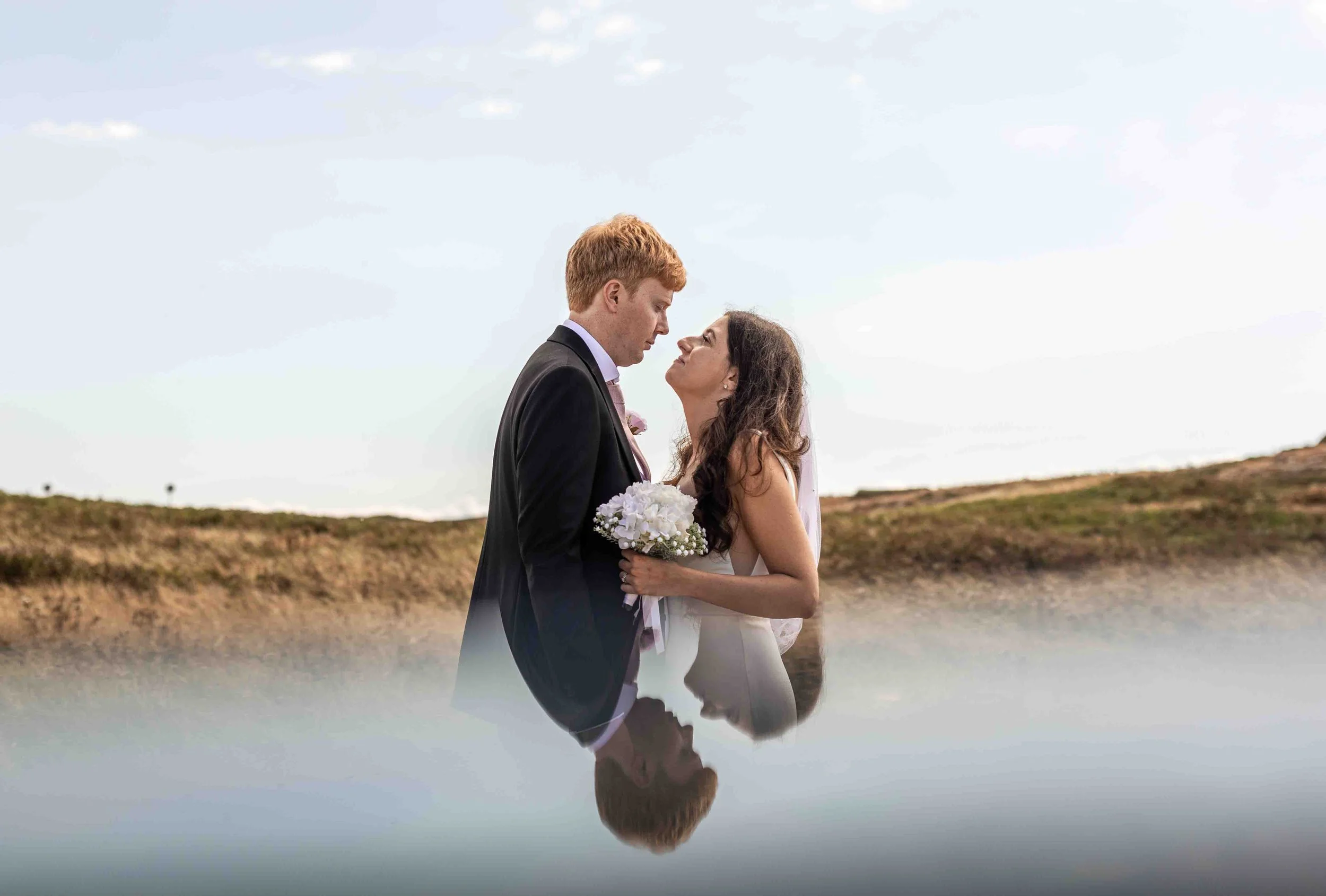 A bride and groom standing close together outdoors with a cloudy sky, holding a bouquet of white flowers, with their reflection visible in a body of water.