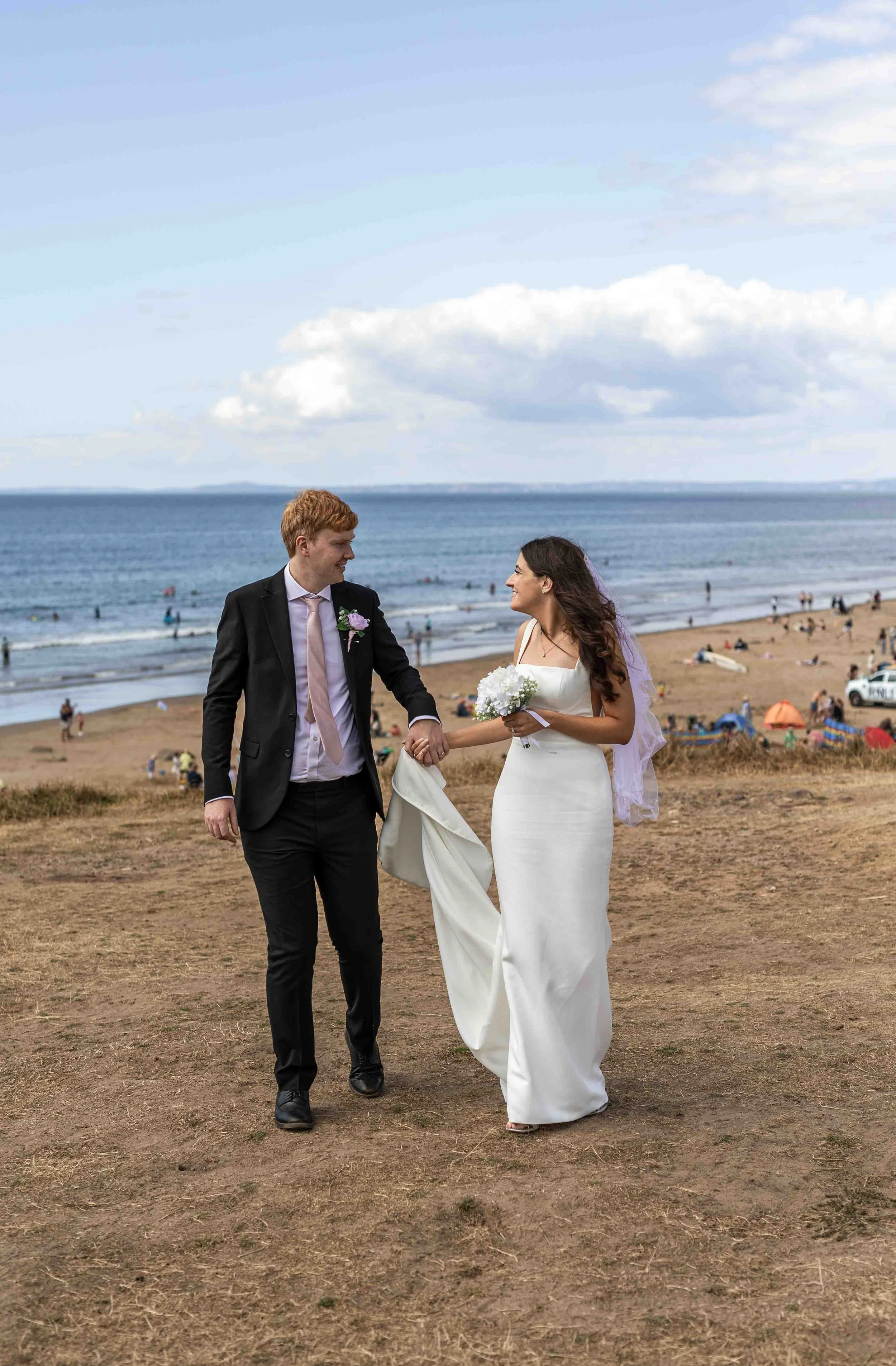 A bride in a white wedding dress holding a bouquet and an orange groom in a black suit with a pink tie walking hand in hand on a beach with sand, ocean, and people in the background.