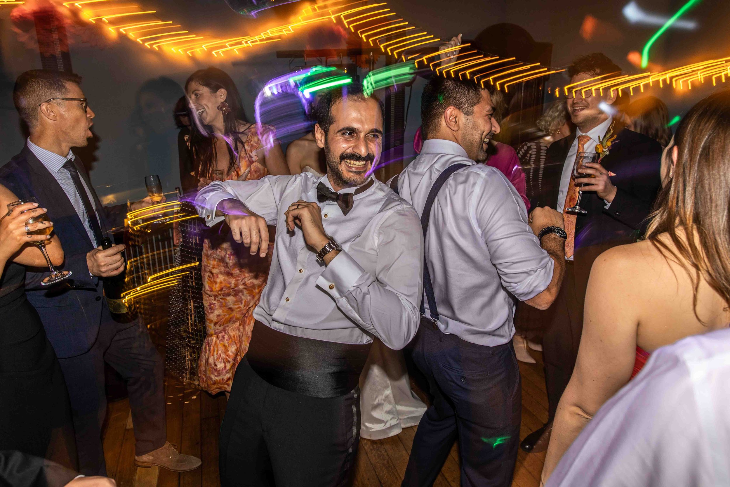 A group of people at a party dancing and having fun, illuminated by colorful lights and wearing formal and semi-formal attire.