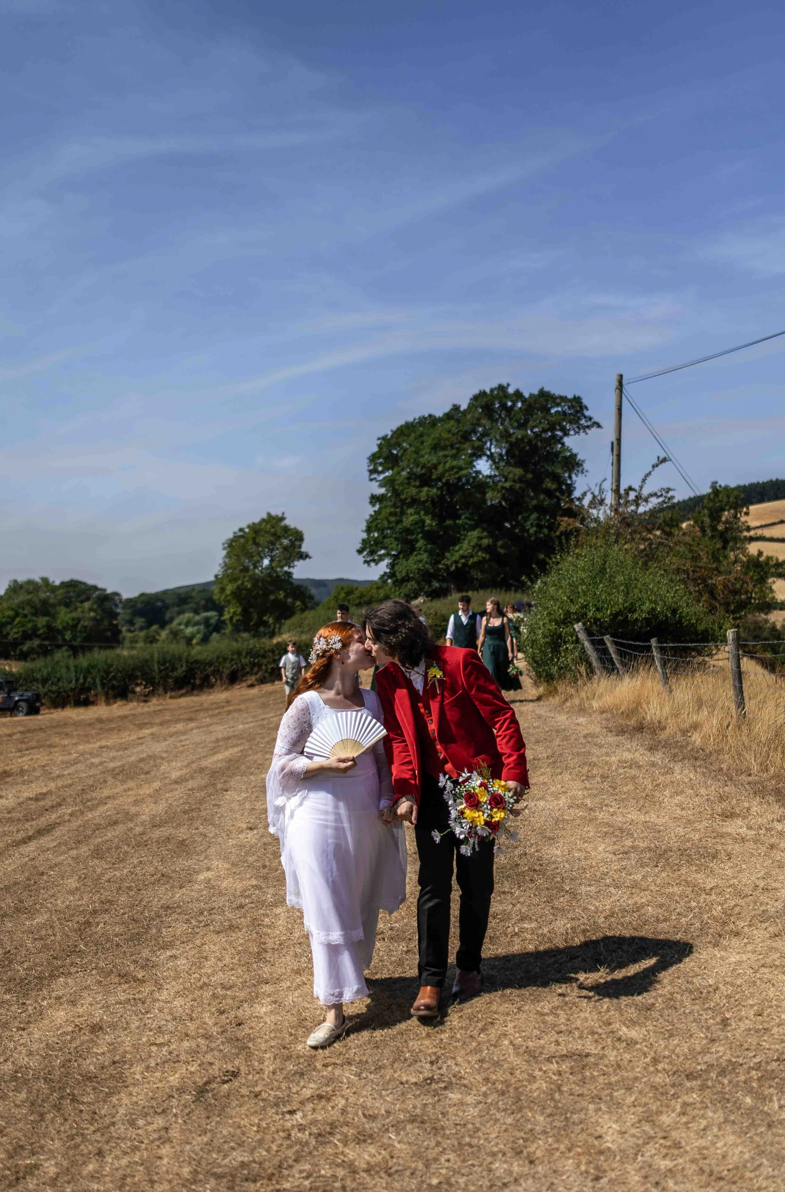 A couple in wedding attire, with the bride holding a fan and a bouquet, walking outdoors on a sunny day with friends or family in the background.