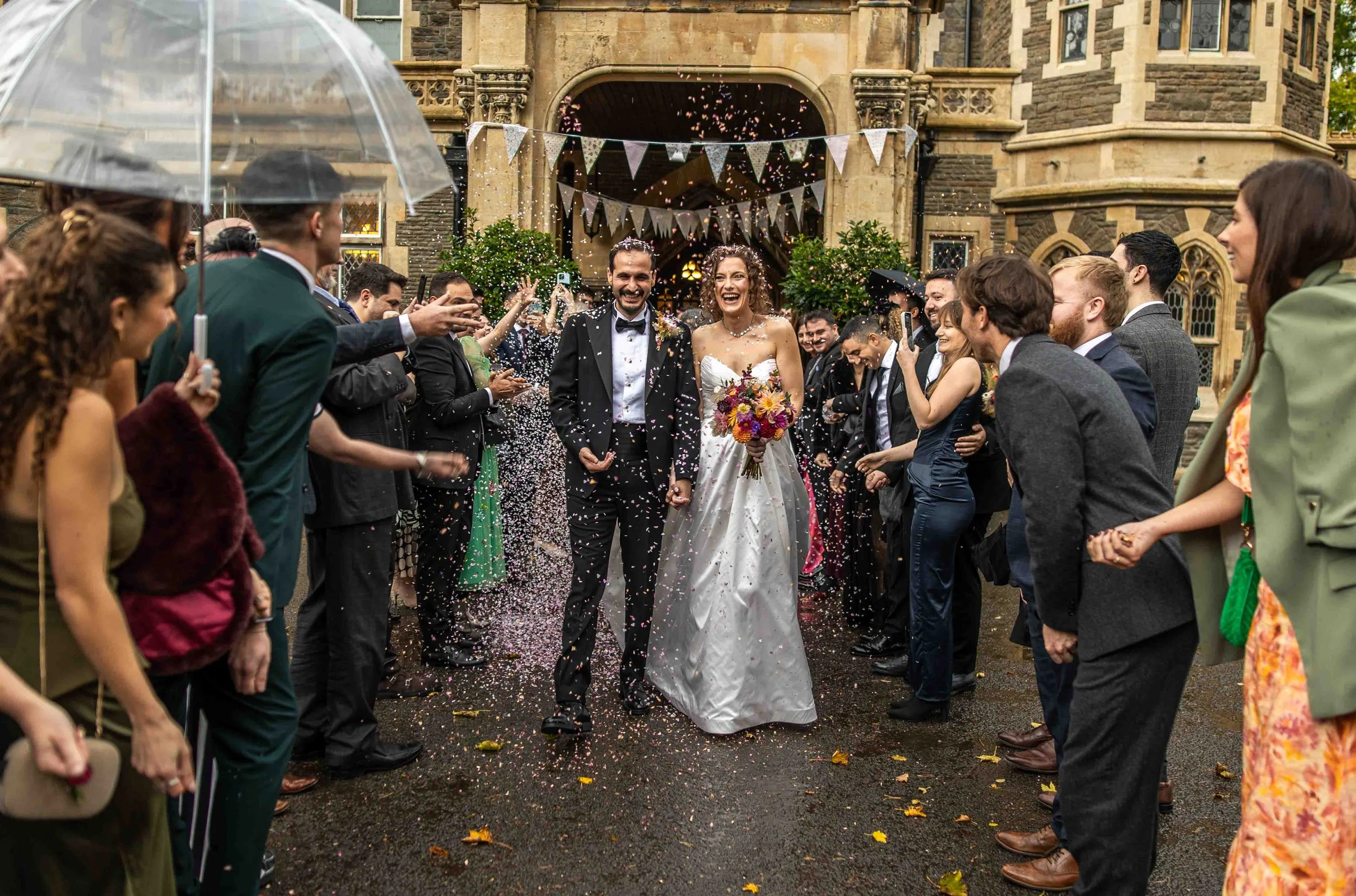 Bride and groom walking hand in hand through a crowd of wedding guests throwing confetti outside a historic stone building.
