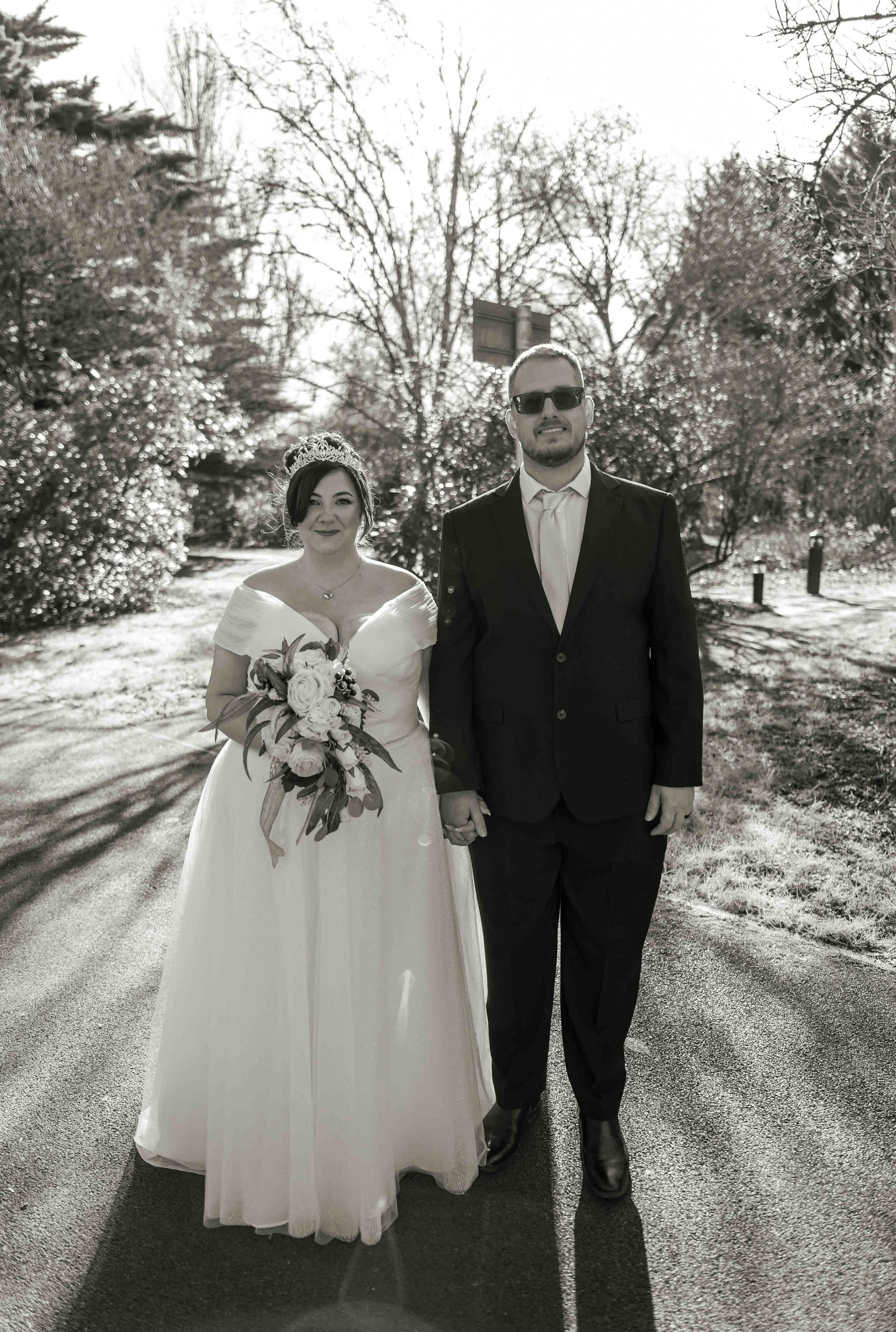 Black and white photo of a bride and groom holding hands, walking outdoors on a sunny day, with leafless trees in the background.