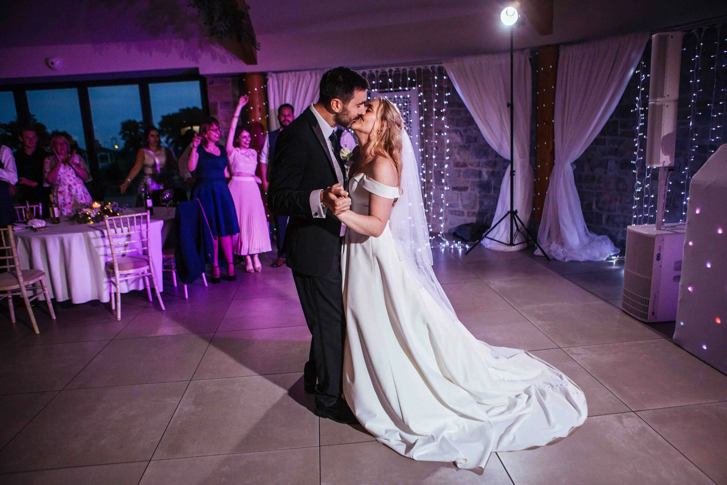Bride and groom sharing their first dance at wedding reception, with guests watching in background, decorated with string lights and curtains. St Tewdrics House. 